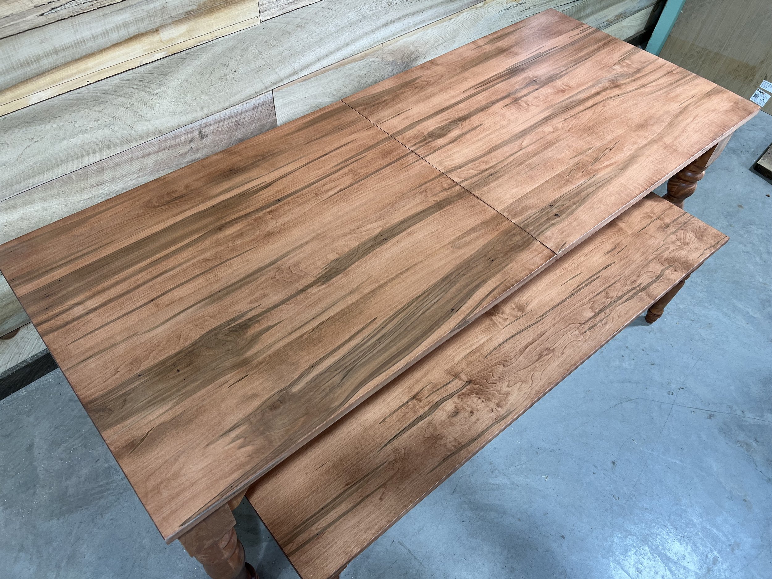 Two wooden tables with a natural finish, one placed on top of the other, in a furniture store or workshop, with concrete flooring and light-colored wooden wall panels in the background.