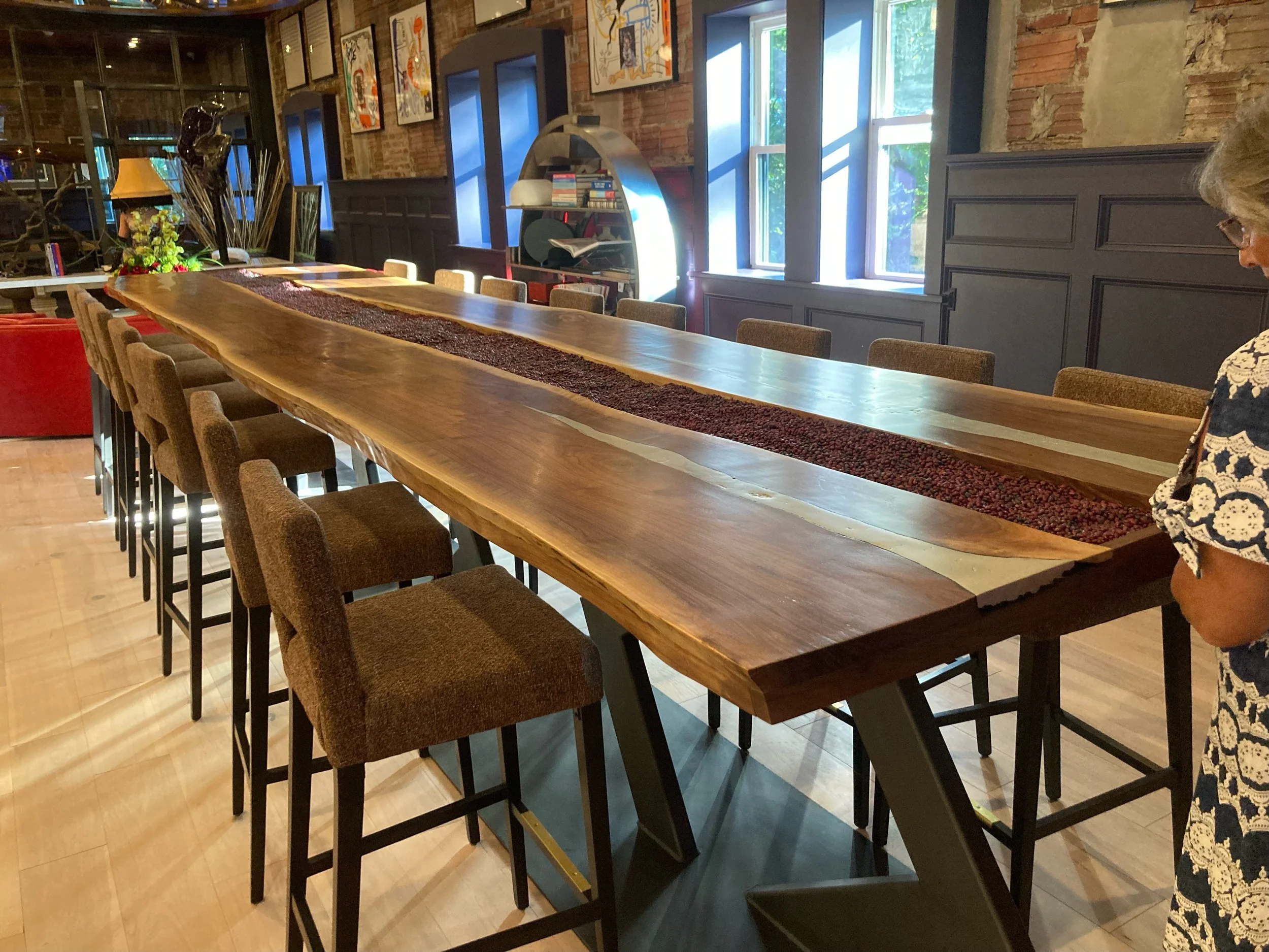 A long wooden table with a red decorative runner in the center, in a rustic-style dining room with exposed brick walls, framed artwork, and large windows.