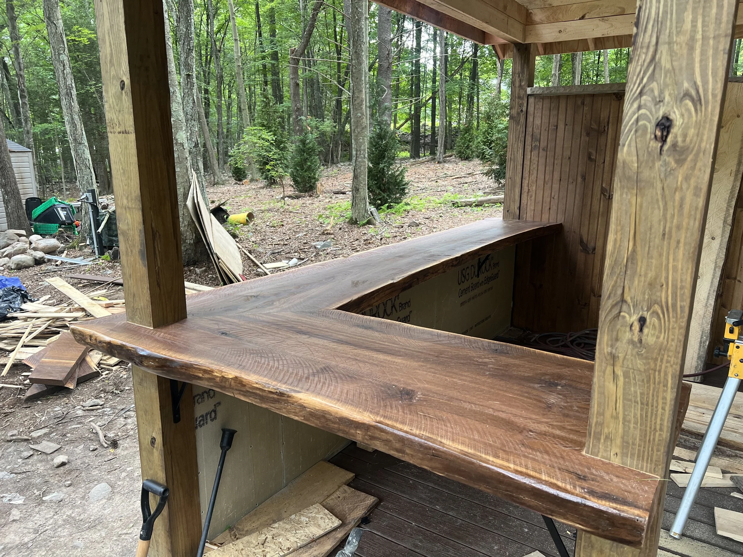 Wooden outdoor bar under construction in a wooded backyard with trees and construction tools visible.