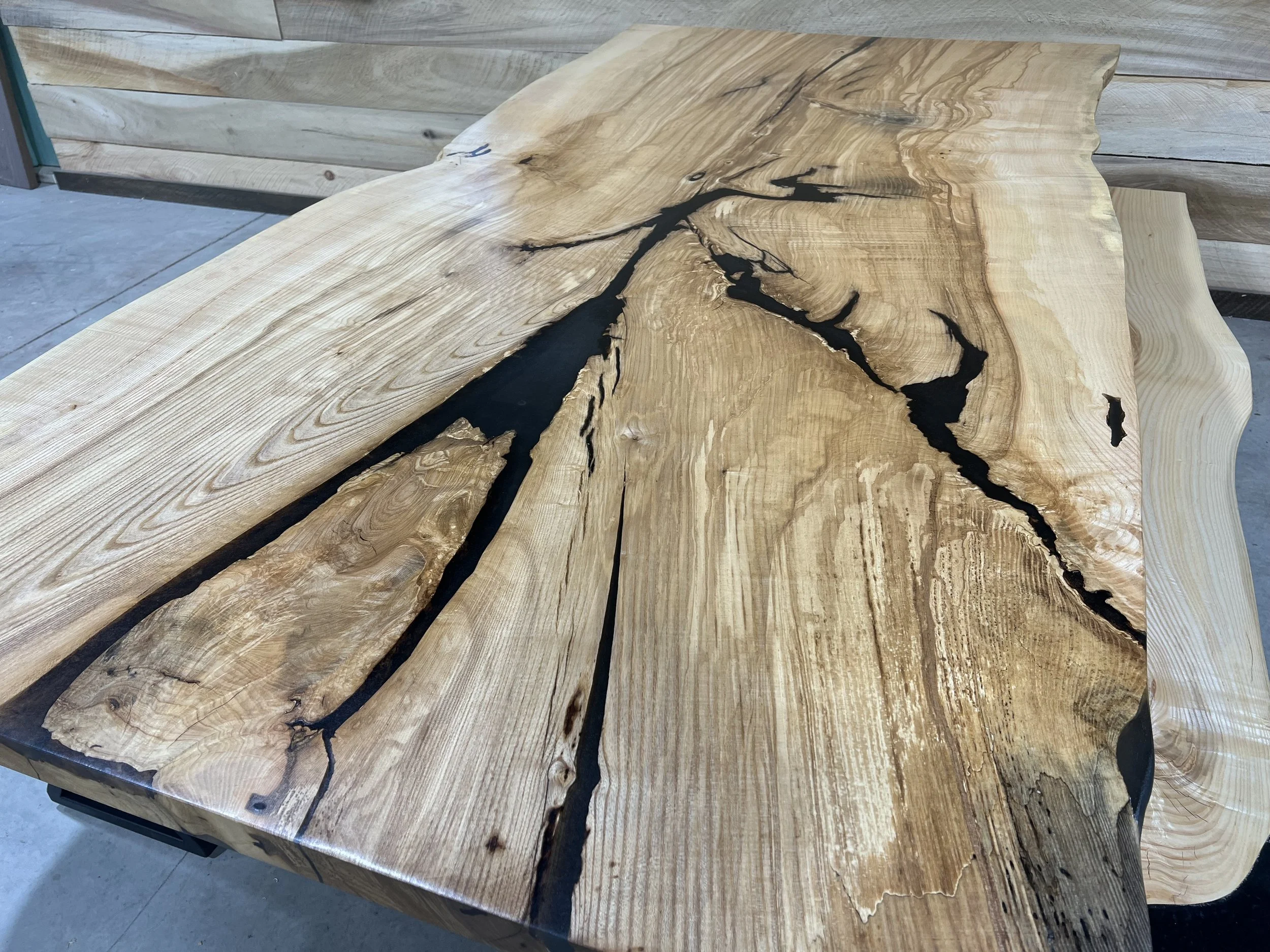 A large, live-edge wooden tabletop with natural grain and cracks filled with black epoxy resin, showcasing a rustic and artistic design.