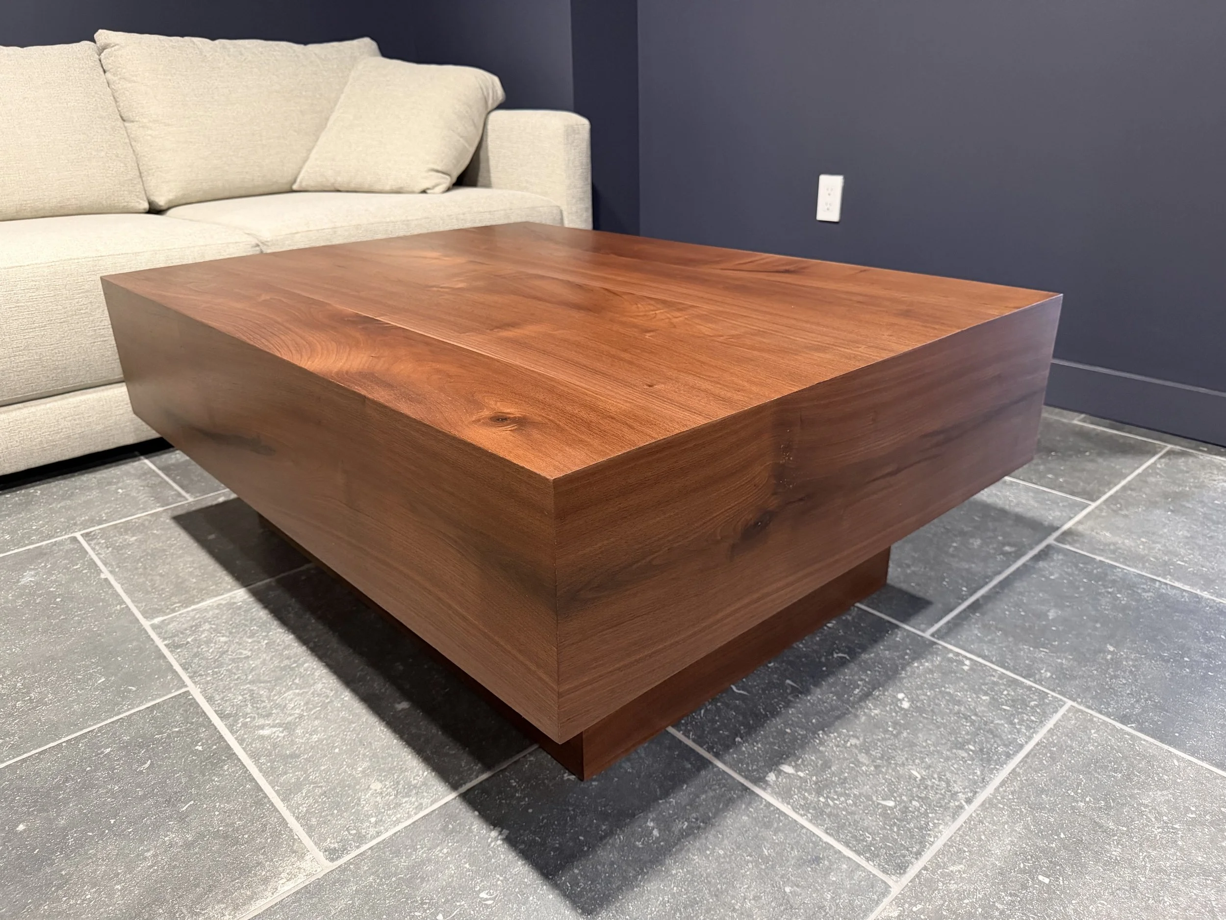 Close-up of a large, rectangular wooden coffee table with a smooth finish, placed on a tiled floor in front of a light-colored sofa and a dark wall.