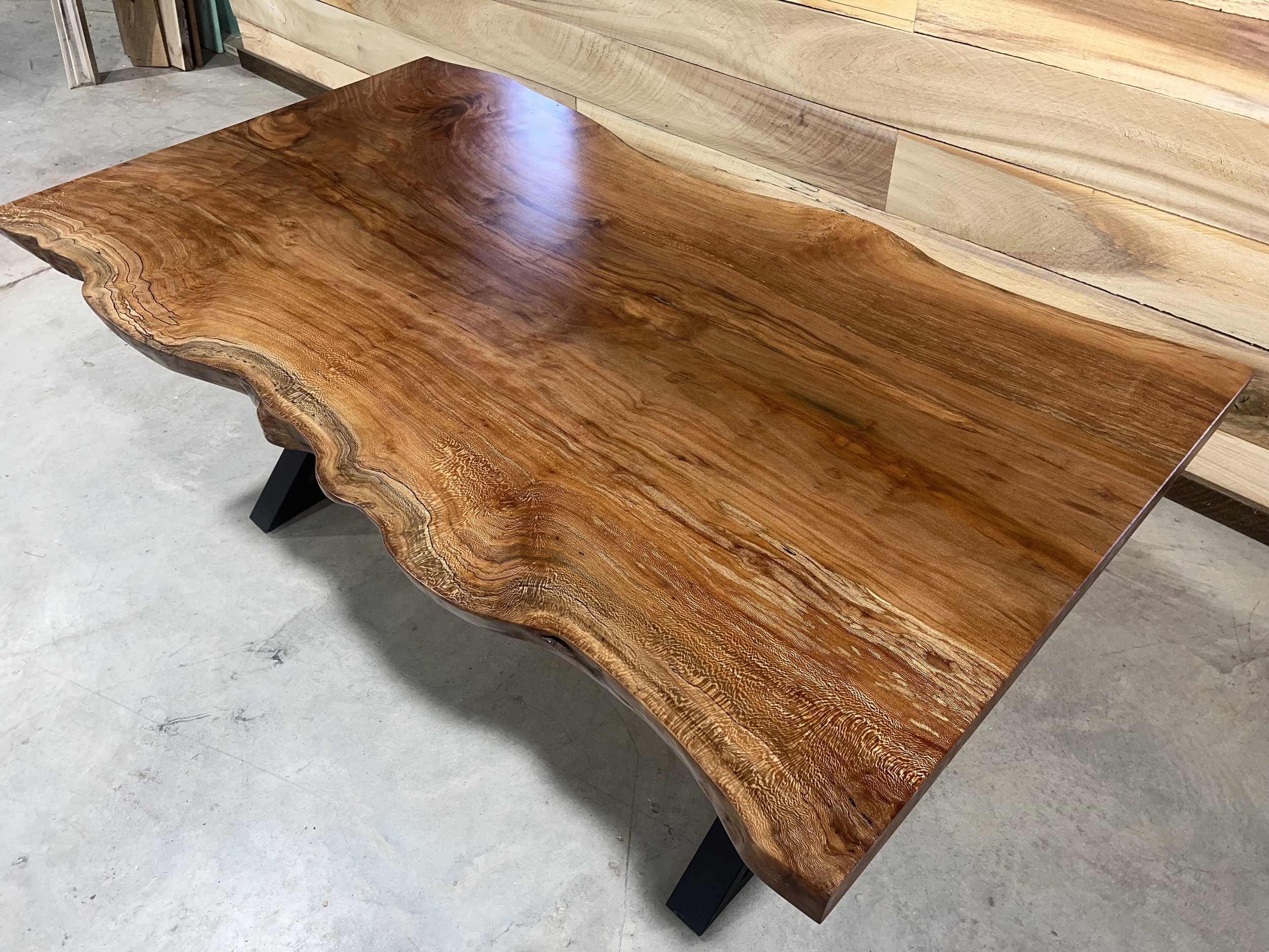 A wooden table with a live edge and a polished surface, sitting on black metal legs, in a workshop or showroom.