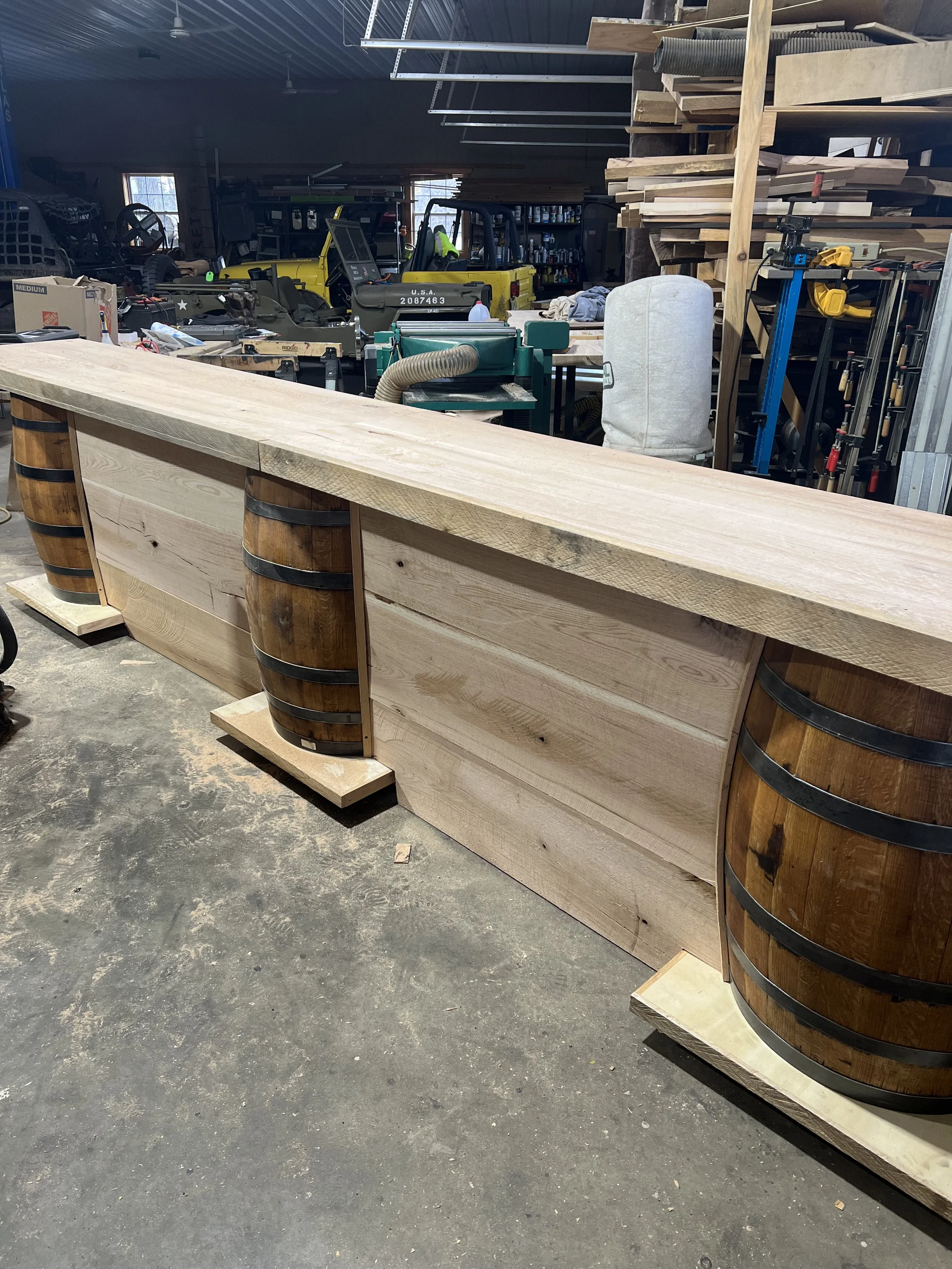 A large wooden bar in a woodworking shop, with a thick, smooth wooden top and barrel-shaped supports on either end, amidst various woodworking tools and machinery.