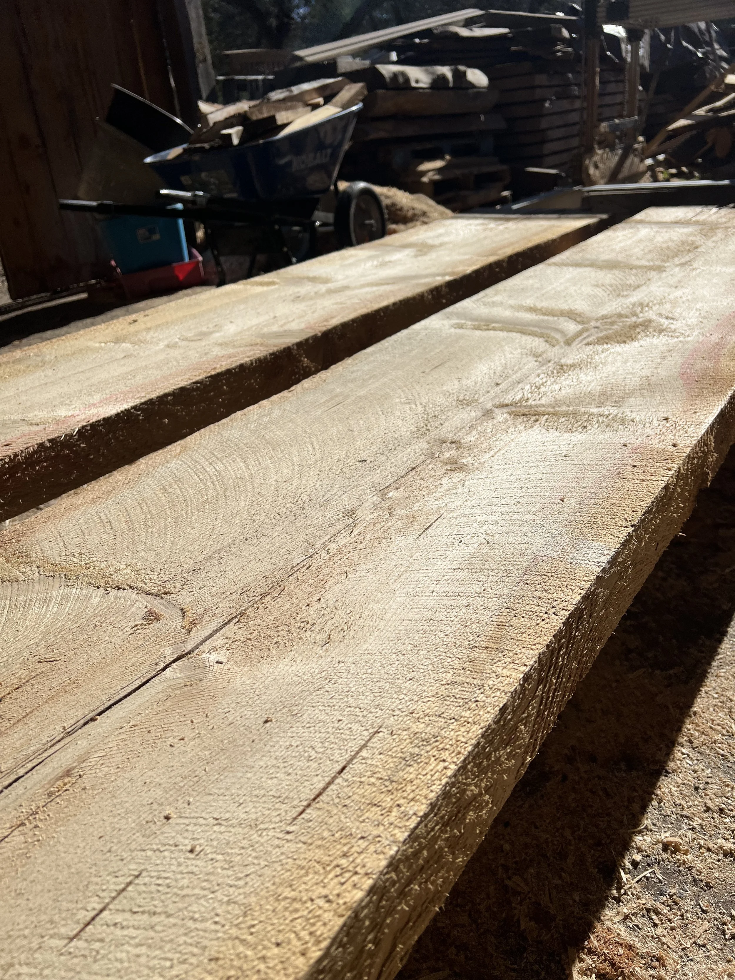Close-up of wooden planks and sawdust in a woodworking yard with stacked lumber and a cart in the background.