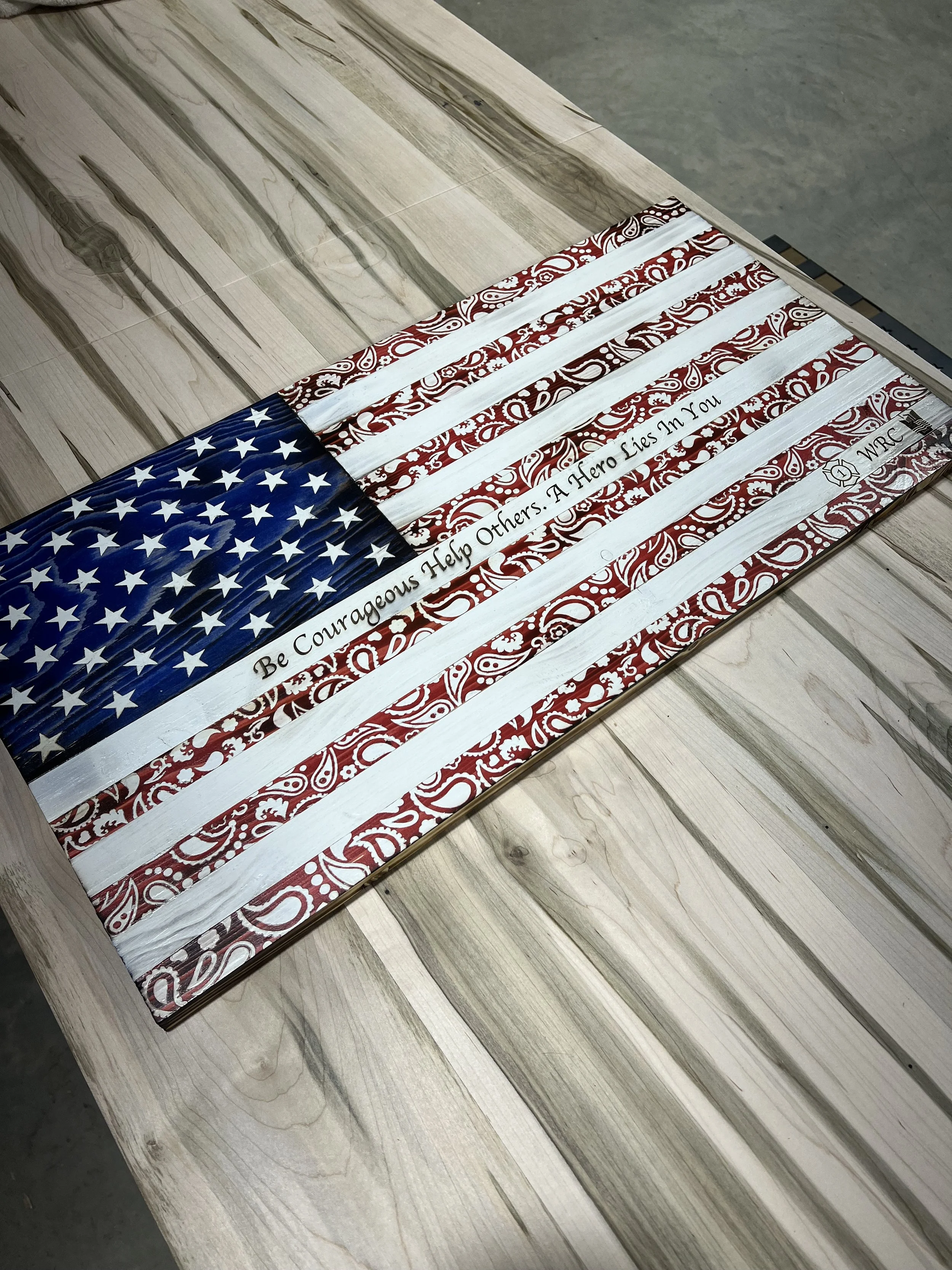 Decorative American flag with a message reading 'Be Courageous Help Others. A Hero Lies In You' on a wooden surface.
