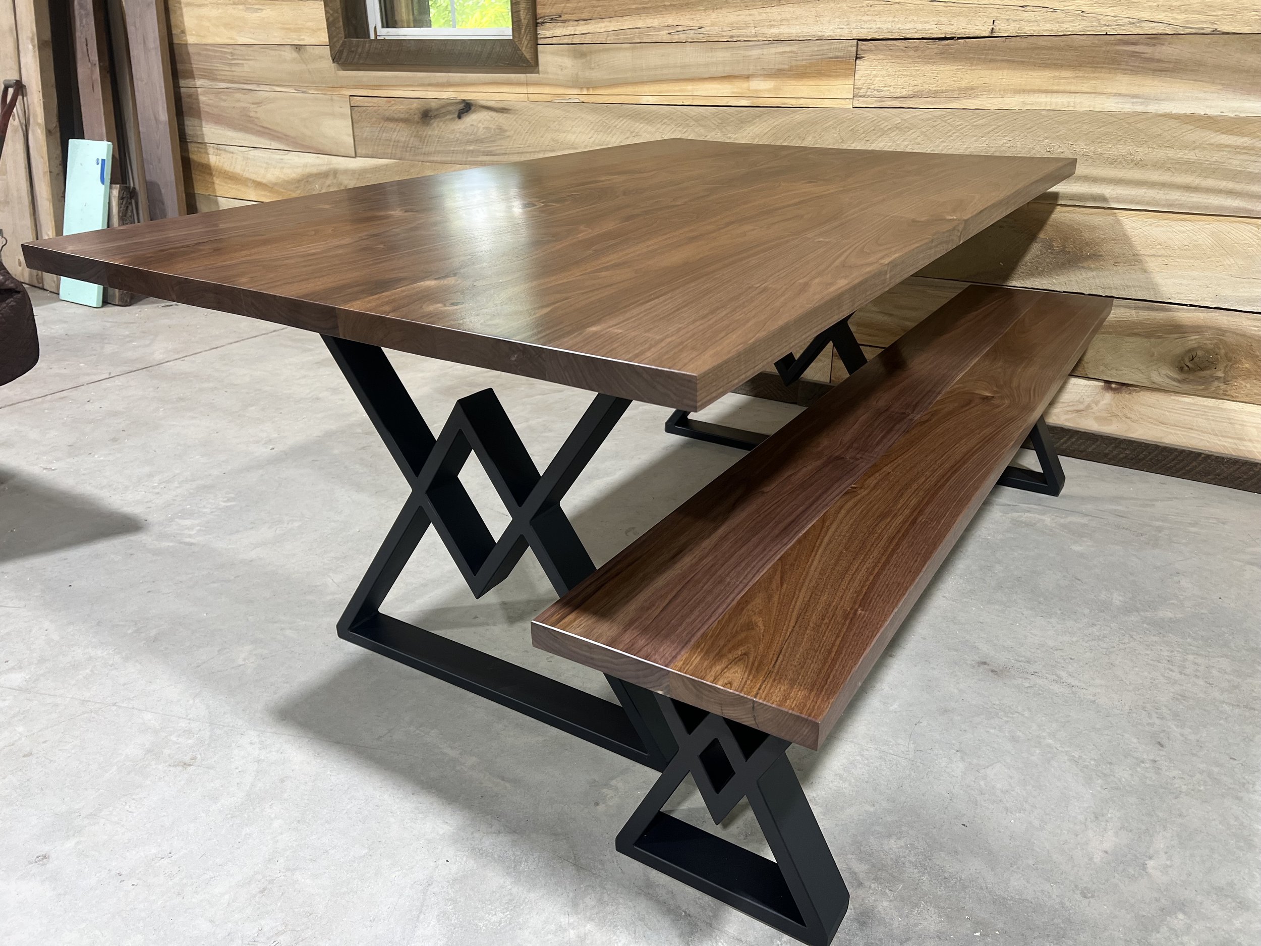 A modern dining table with a thick wooden top and black metal legs, accompanied by a matching wooden bench, set against a wooden wall background in a workshop or furniture store.