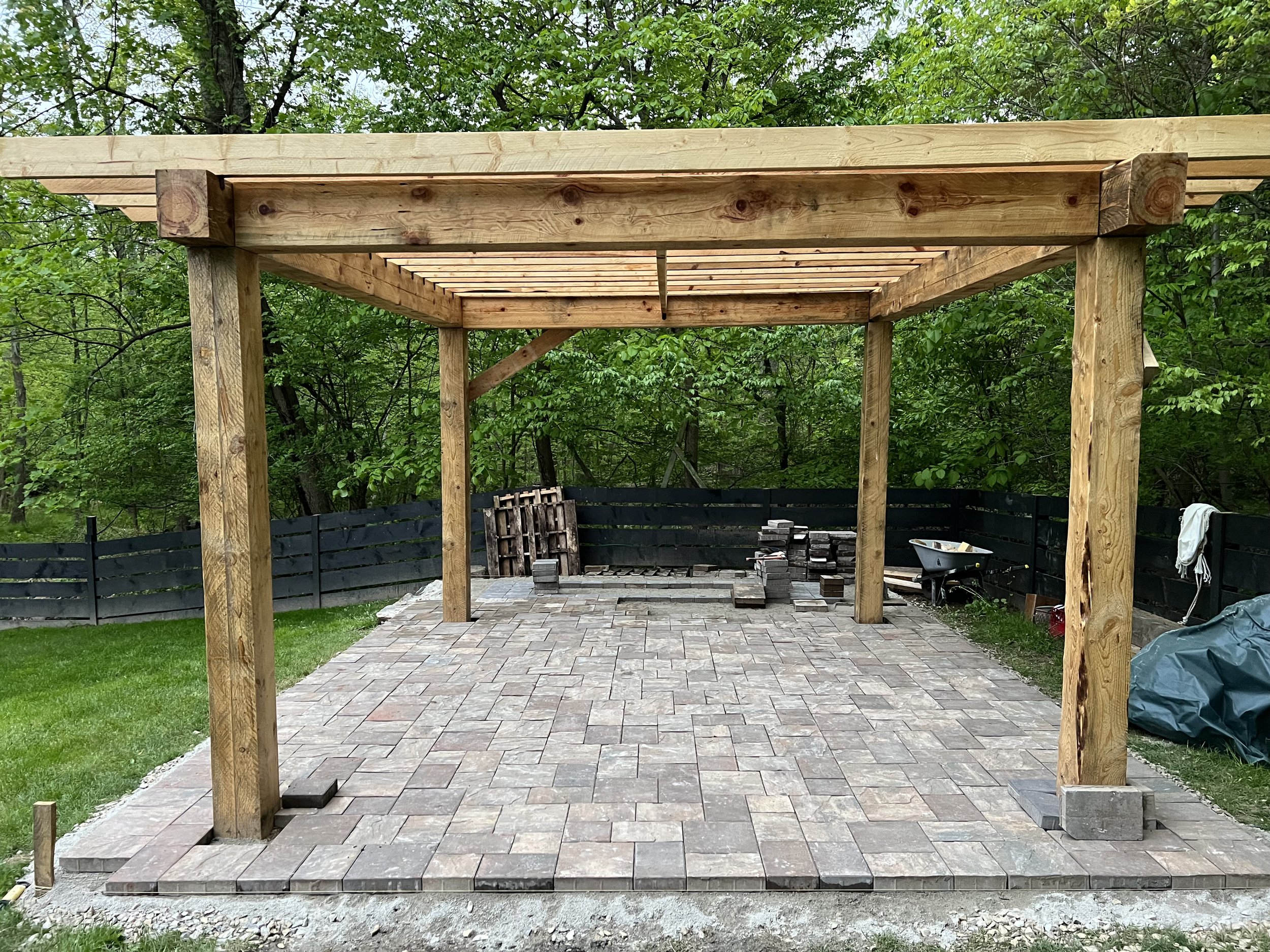 A partially constructed outdoor patio with a wooden pergola, paved with interlocking stone bricks, in a backyard with green trees and a black fence.