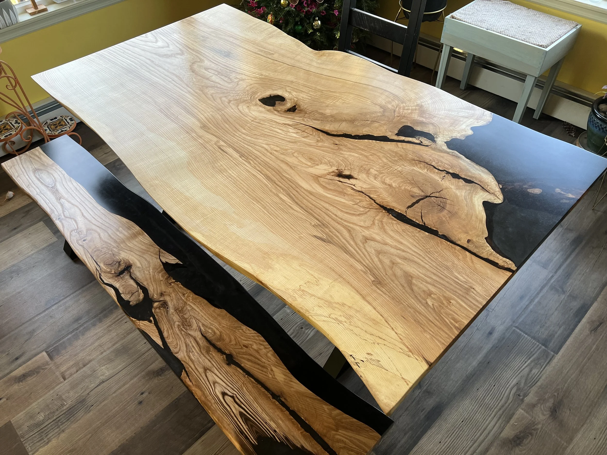A large wooden dining table with natural wood grain and black resin inlays, matching benches, and a holiday tree in the background with ornaments.