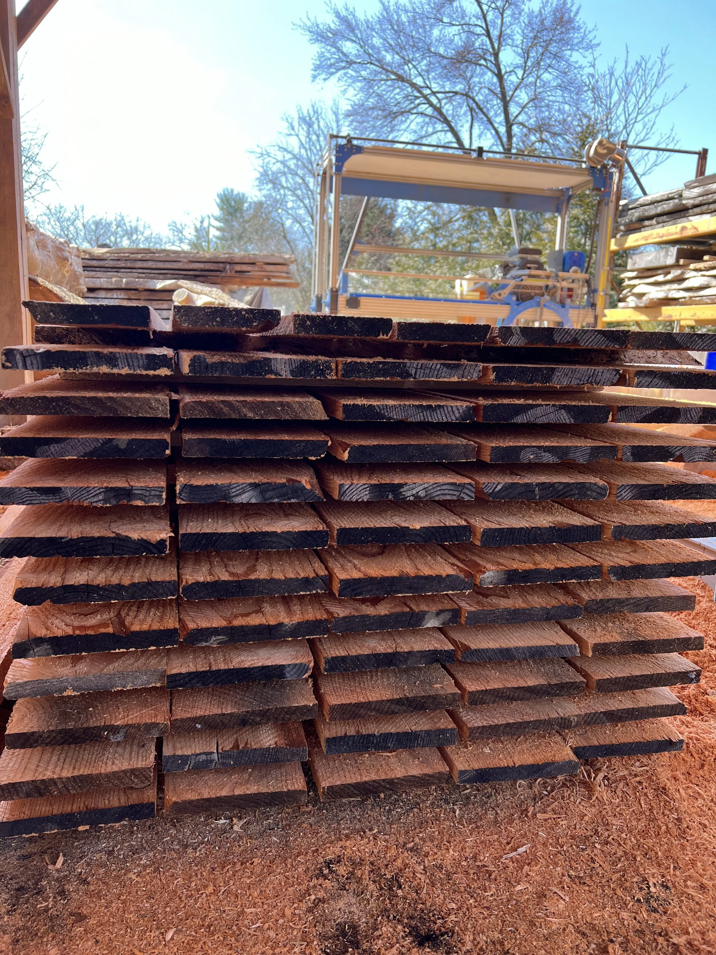 Stacked cut wooden planks in a lumber yard with woodworking equipment in the background and trees in the distance.