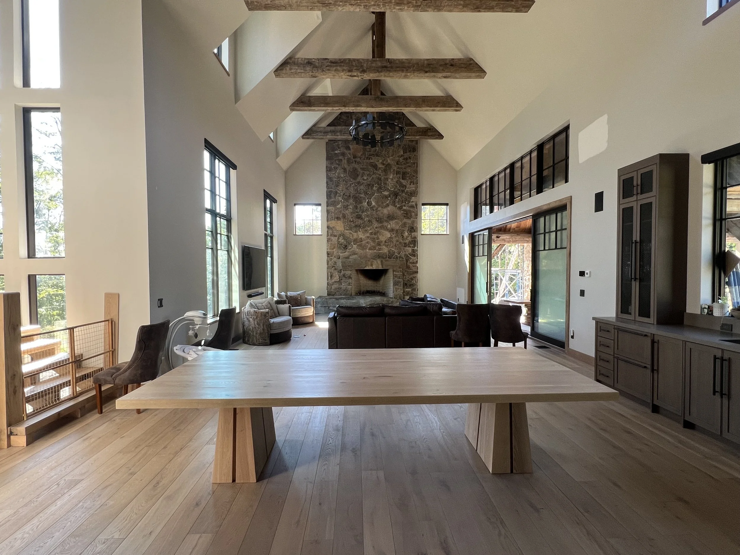 Open-concept living room with high vaulted ceilings, large windows, and exposed wooden beams. There's a stone fireplace, a sectional sofa, and various chairs. Natural light fills the space, which features light wood flooring and modern furnishings.