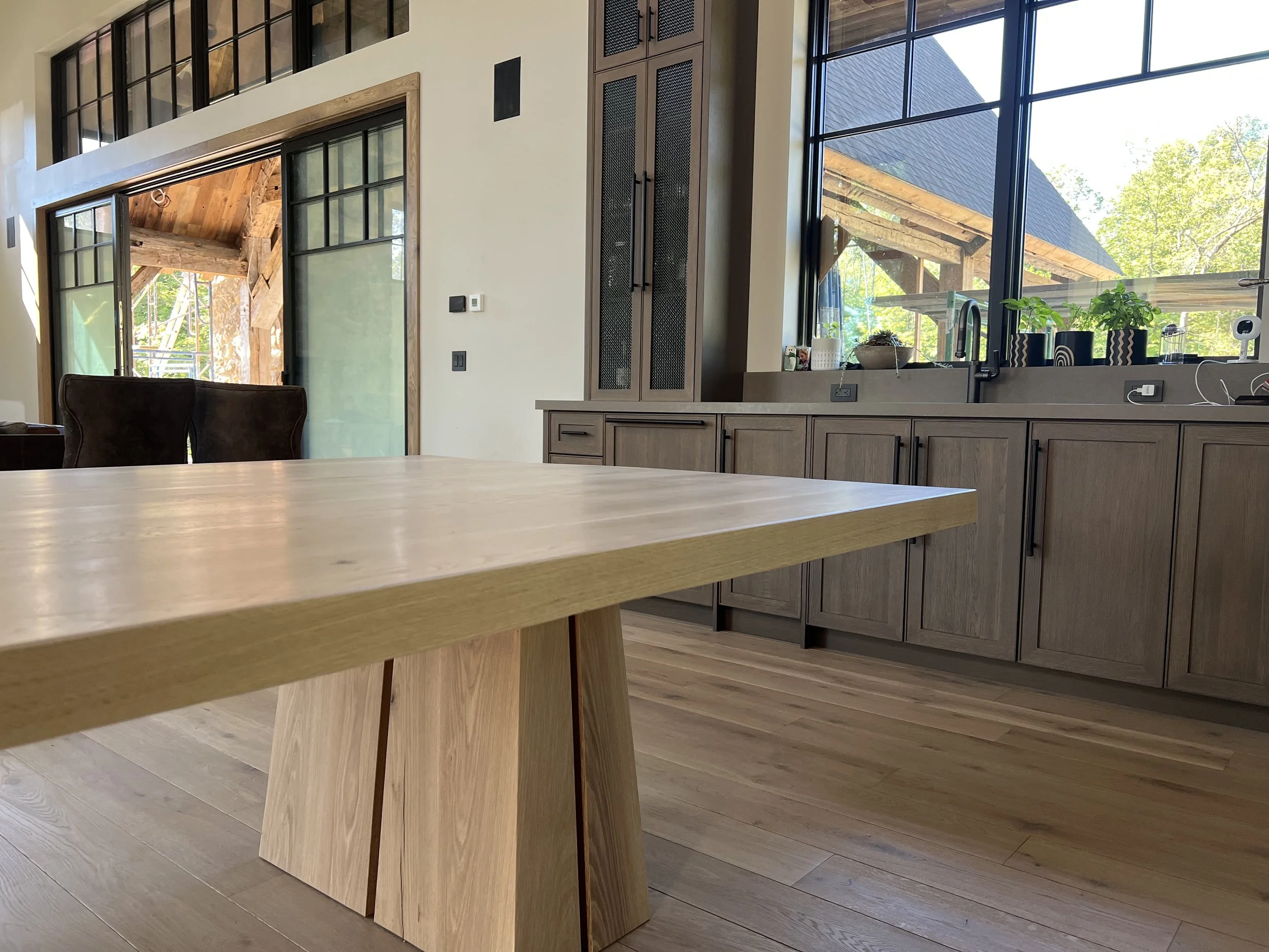 Modern kitchen with large windows, wooden cabinetry, and a wooden dining table with a distinctive angled leg design. Outside, a wooden structure is visible through the windows, with trees and sunlight indicating a bright day.