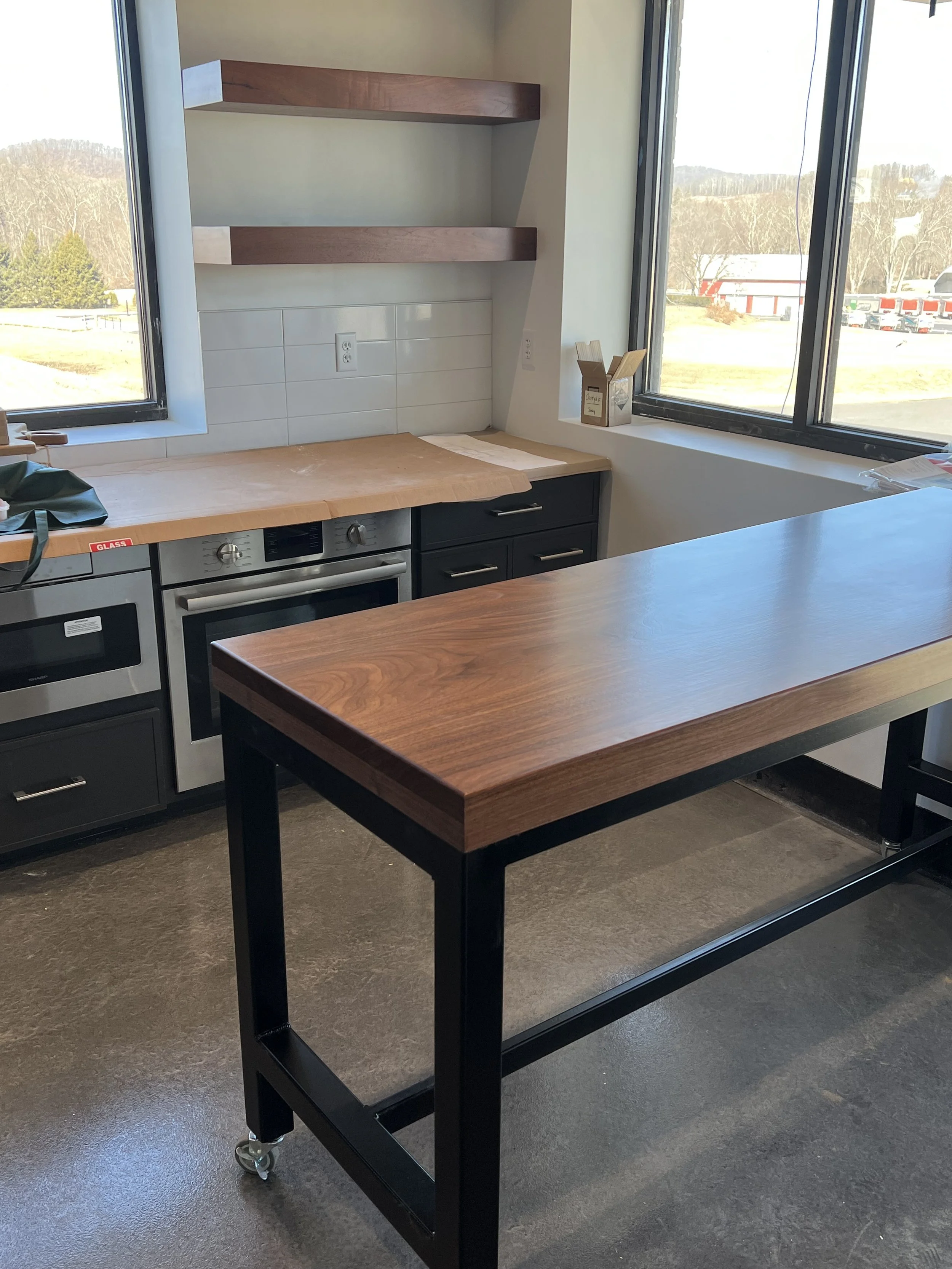 Modern kitchen with black cabinetry, stainless steel oven, wooden countertop, empty wooden table with black metal frame on casters, large windows showing outdoor view, white tile backsplash, small wooden shelves, and a paper towel holder.