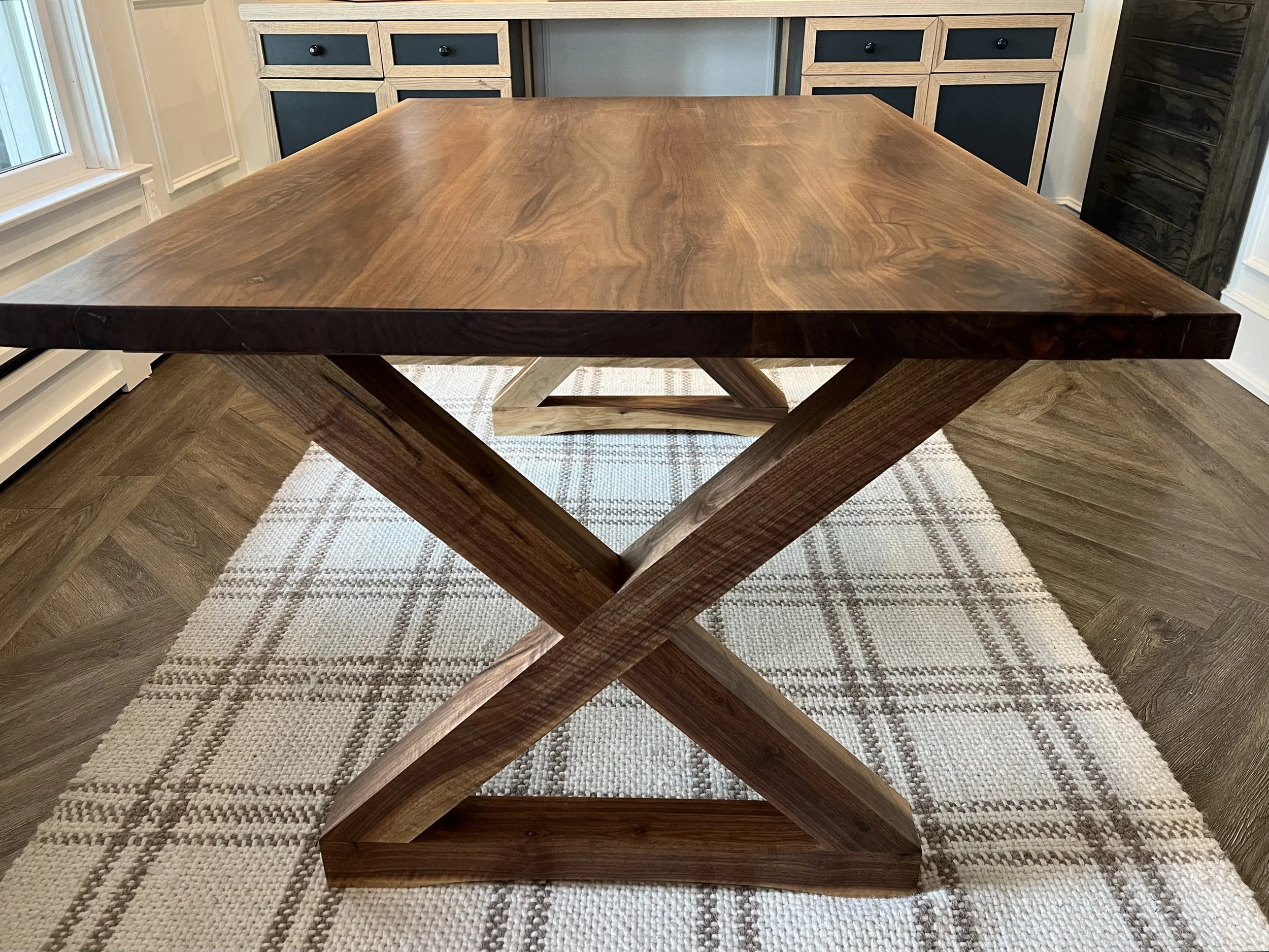 A wooden dining table with a rectangular top and crisscrossed legs, placed on a beige and brown plaid rug in a room with wooden flooring and dark-colored cabinet doors in the background.