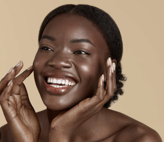 A smiling Black woman with short, natural hair touching her face with both hands against a beige background.