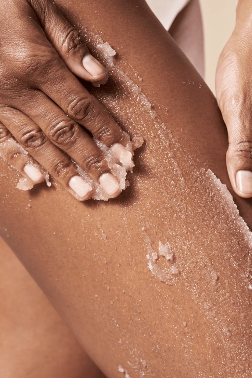 A person applying a granular exfoliating scrub onto their leg, with one hand spreading the scrub and the other hand gently rubbing it in.