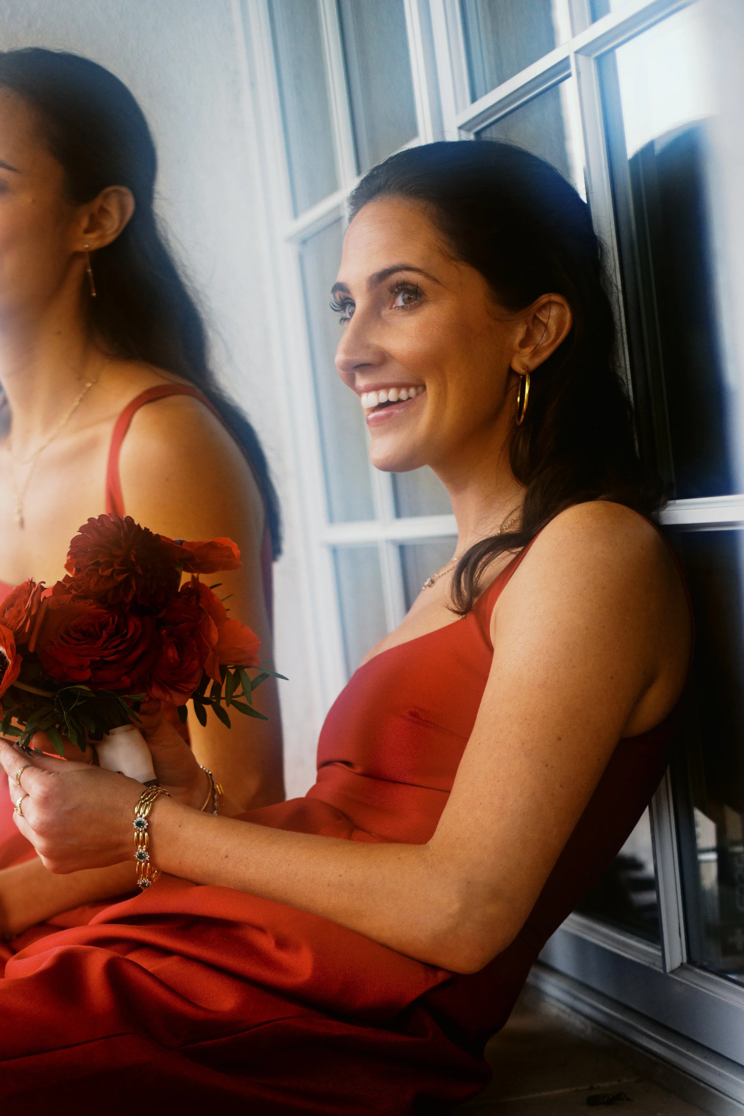 A bridesmaid sits outside during a break. She is wearing deep red and holding a bouquet of red flowers. Photograph taken by wedding photographer Sheridan Mangold at Congressional Country Club in Bethesda, MD.