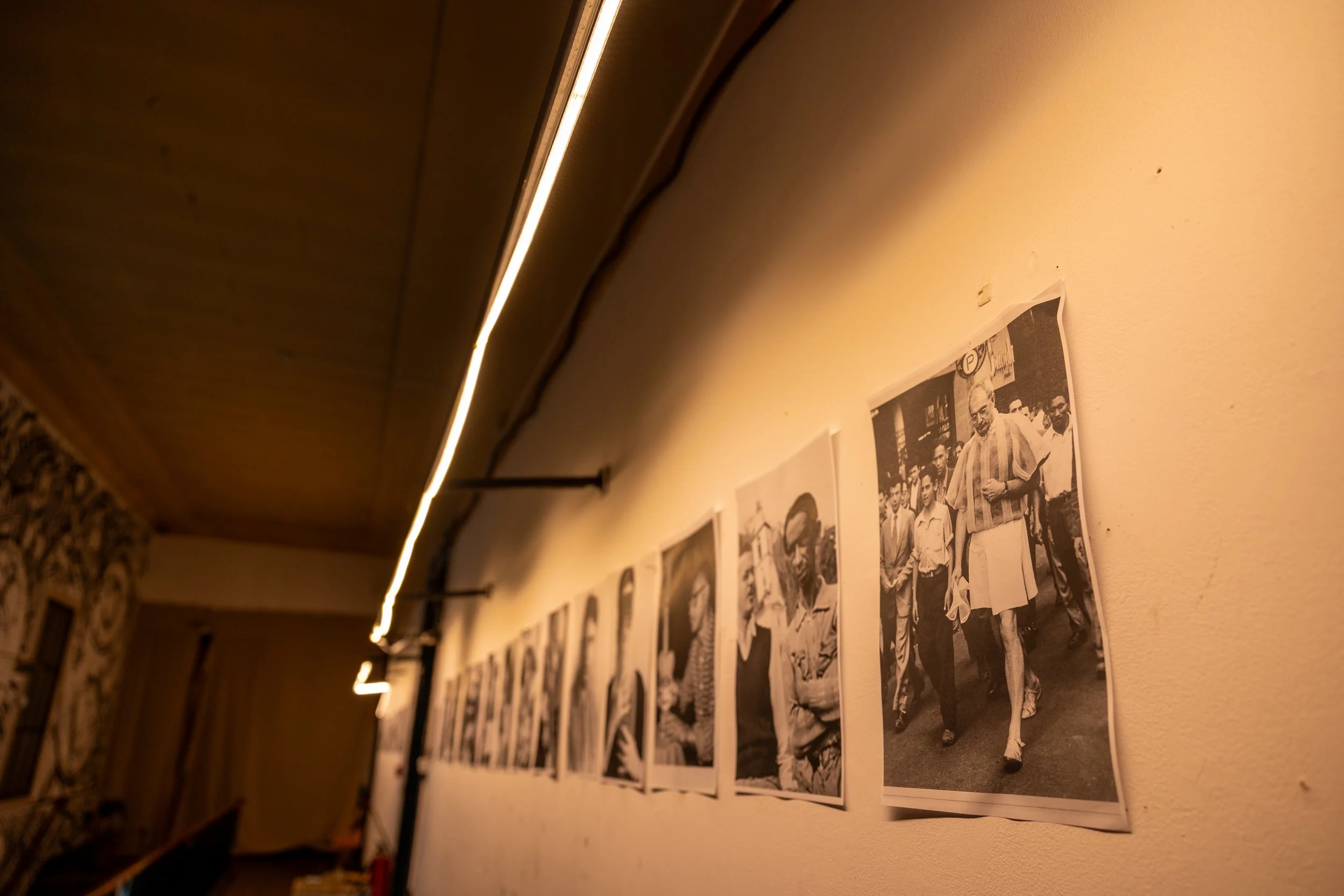 Black and white historical photograph of a group of people, displayed on a beige gallery wall, illuminated by a long, narrow ceiling light.