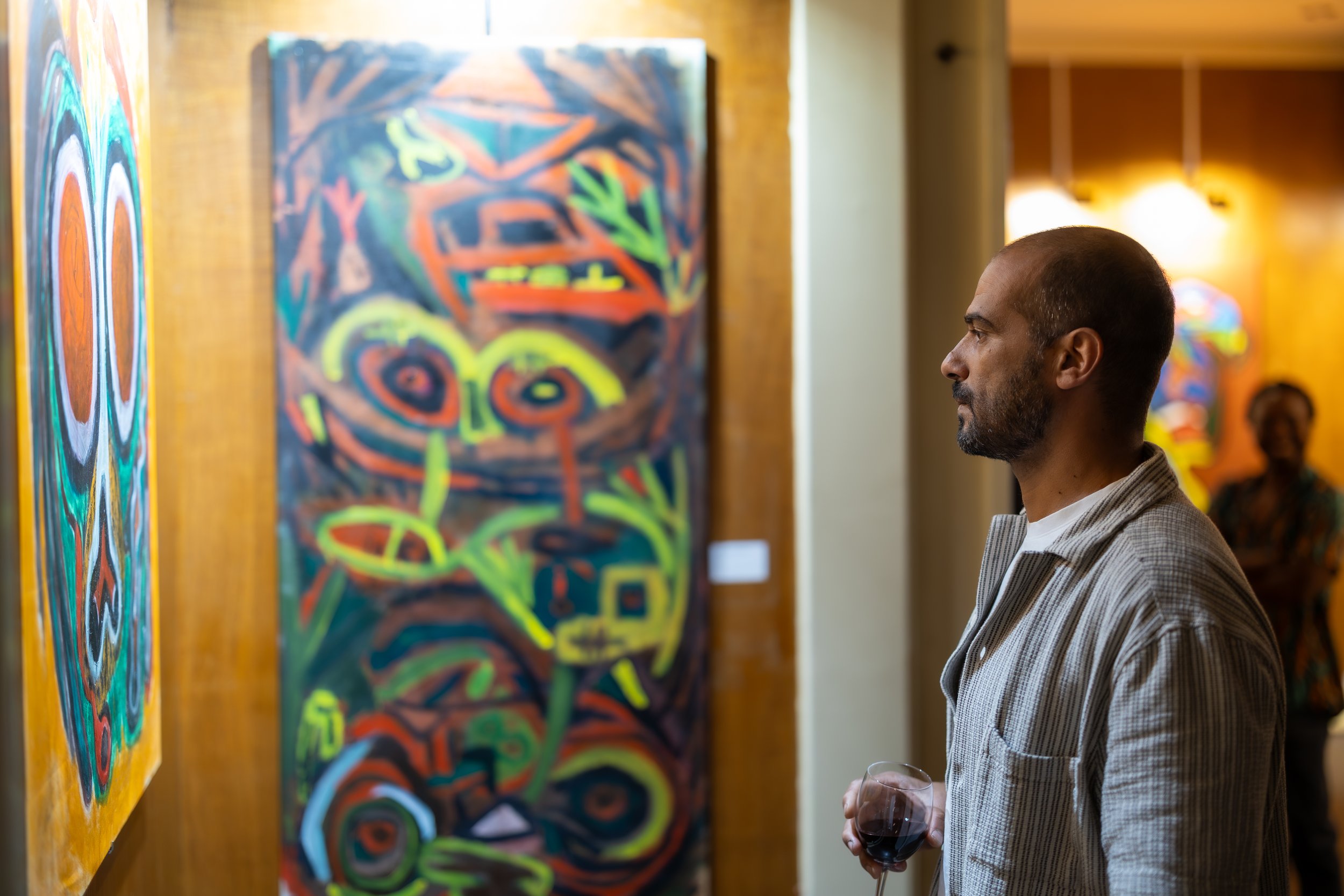 Man with tattoos sitting on a chair in an art studio surrounded by colorful abstract paintings, with art supplies and equipment on the floor and tables.