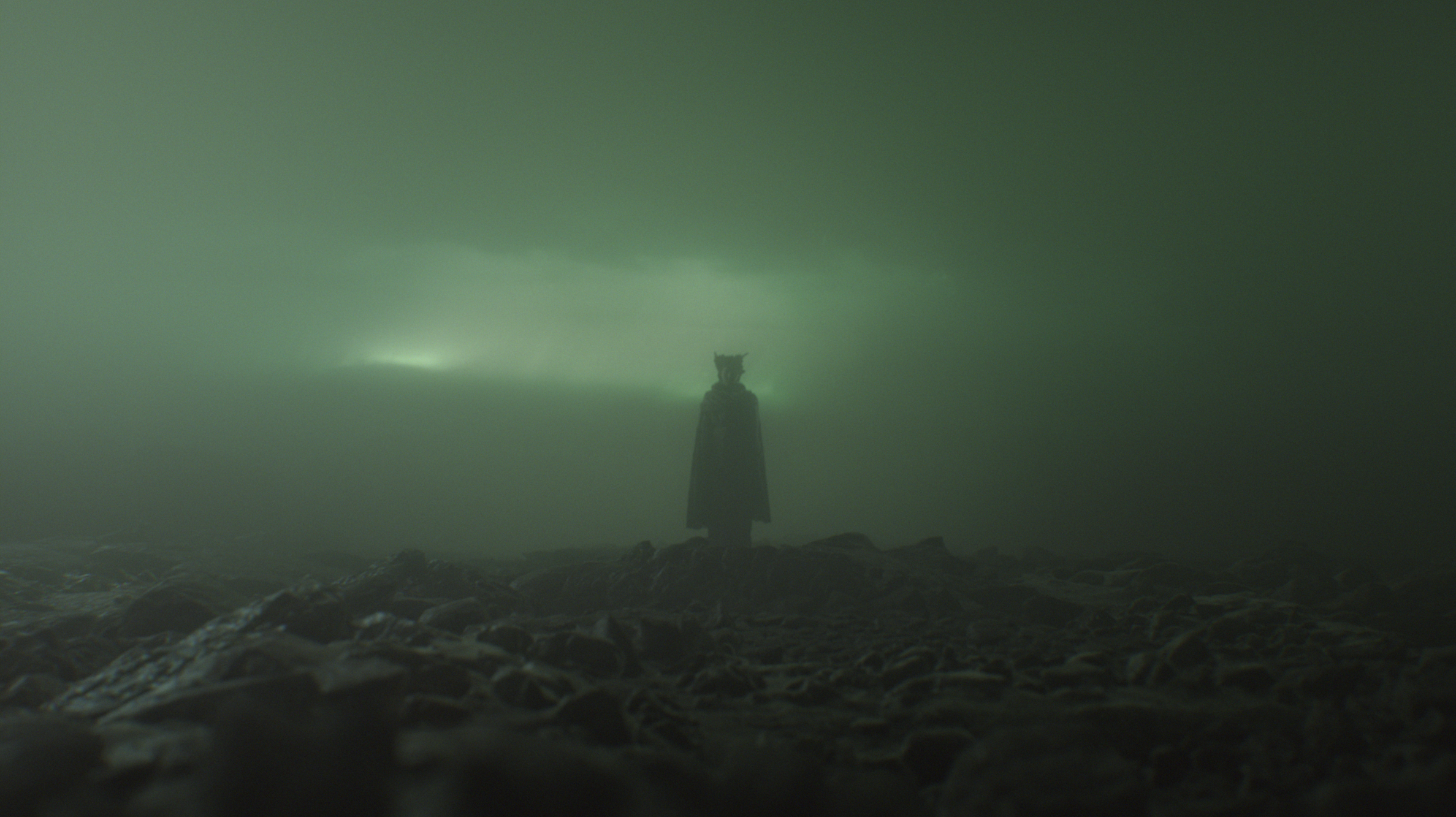 A person dressed in dark clothing and wearing a cat-like mask stands alone on a rocky shoreline in foggy, greenish light.