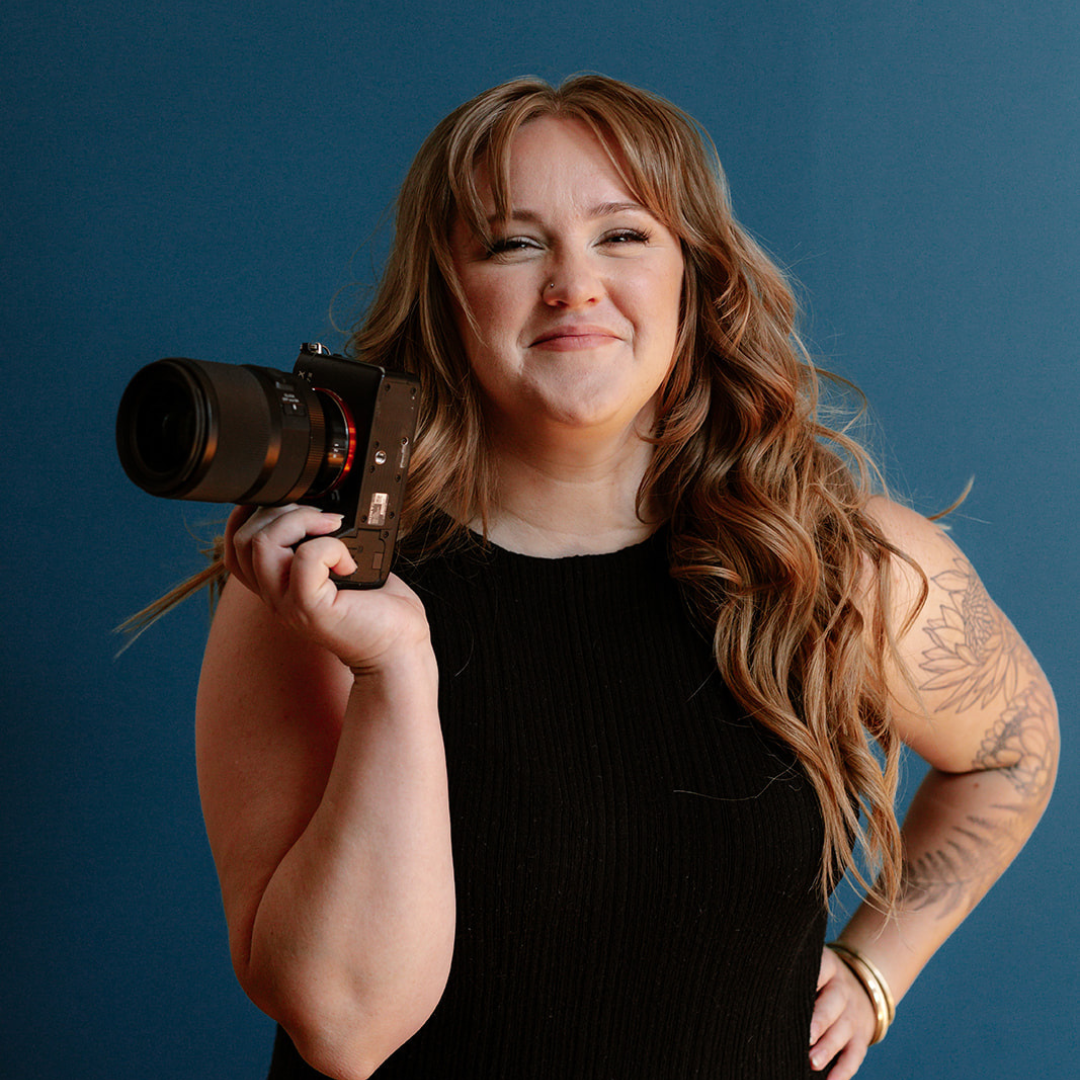 Rachel File, a photographer with long wavy hair holding a camera, smiling, wearing a black sleeveless top, standing against a blue background.