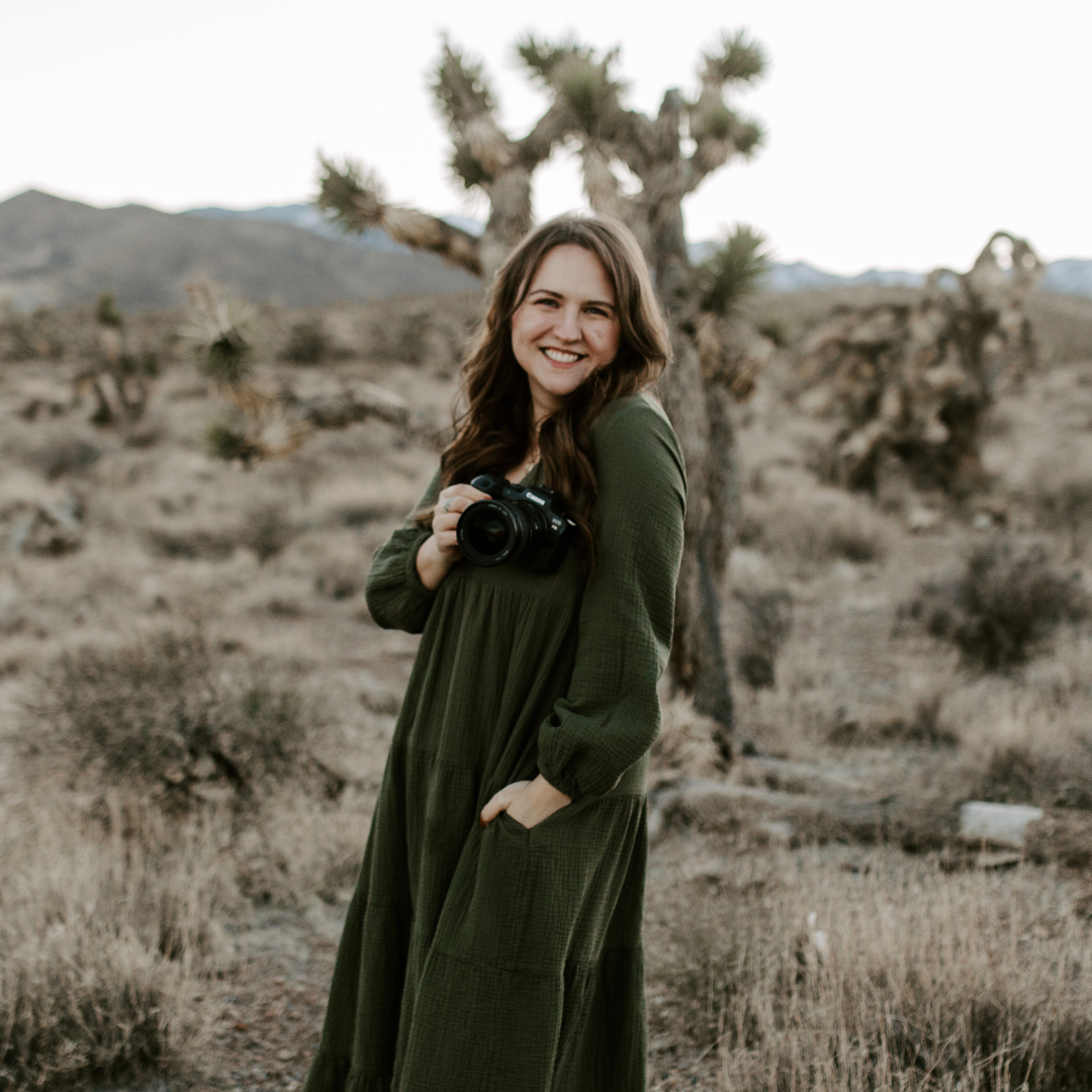 Katelyn Kyles, Photographer smiling in a desert landscape holding a camera, with Joshua trees and mountains in the background.