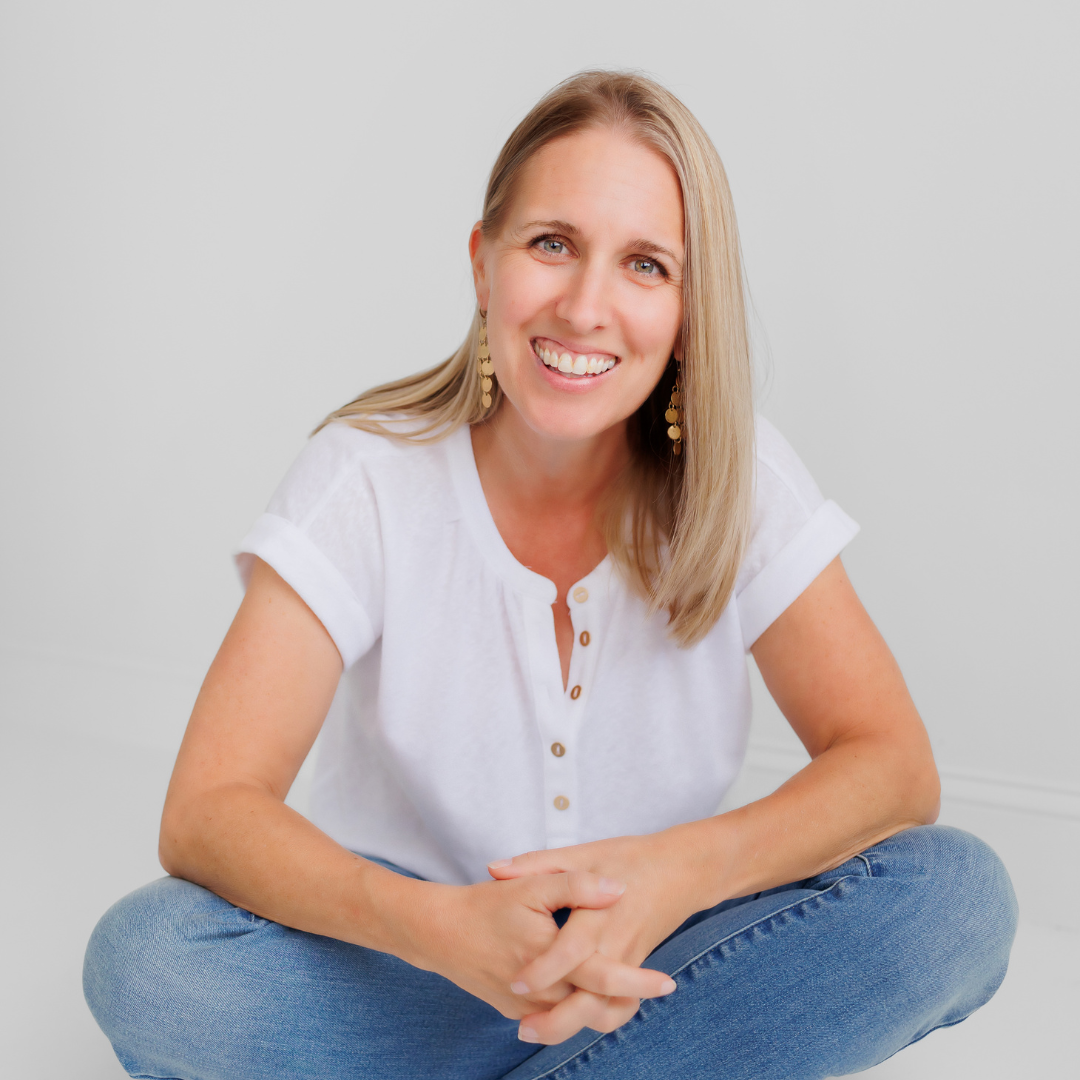 Tara Stuart, organiser. A woman with blonde hair and blue eyes smiling, sitting cross-legged in a white t-shirt and jeans against a plain background.