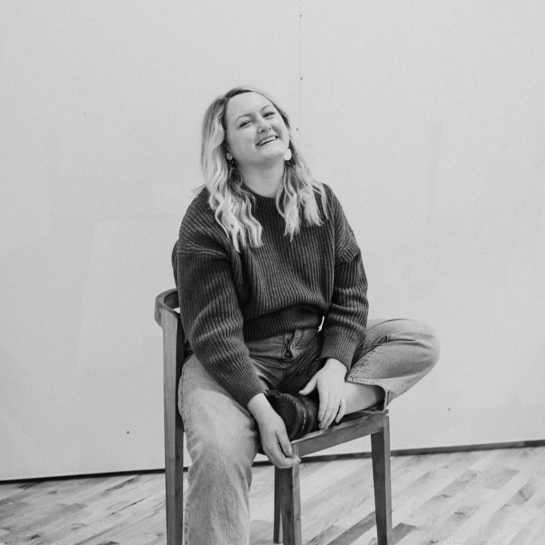 Sidney Scheinberg A woman with long wavy hair sitting cross-legged on a wooden chair, smiling at the camera, wearing a cozy sweater and jeans, against a plain wall background.