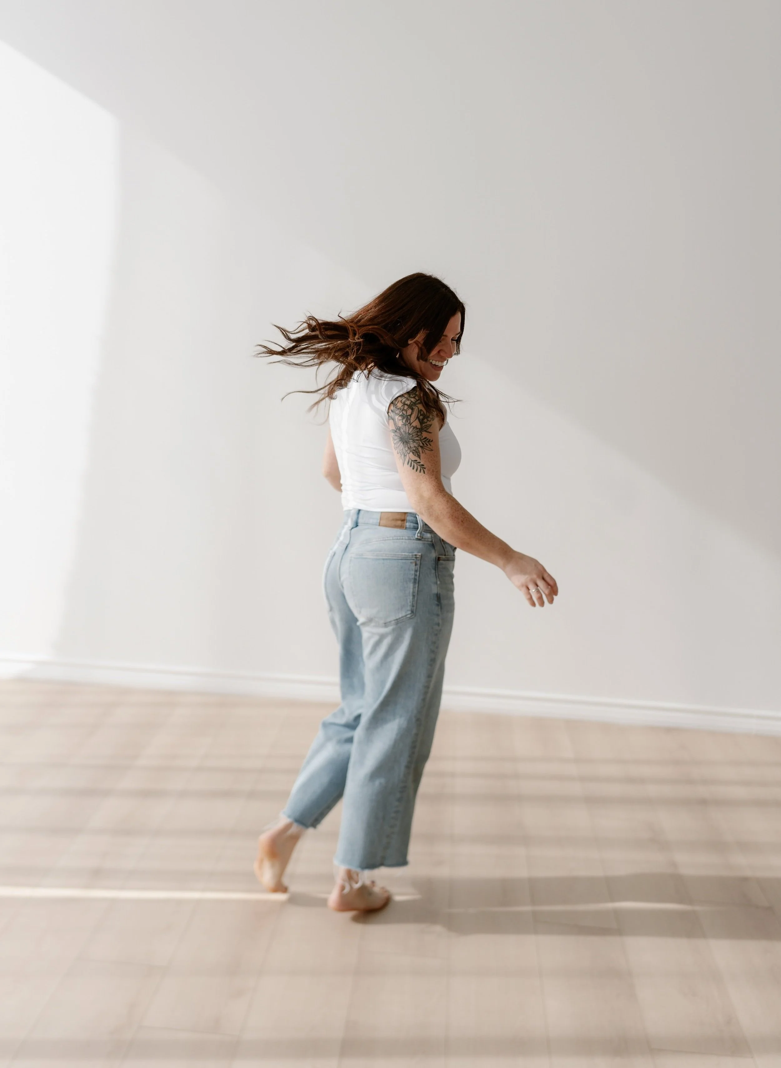 A woman with long brown hair, wearing a white sleeveless top and light blue jeans, is dancing barefoot on a wooden floor in a minimalist, white room. She is smiling, her hair is flowing, and she has a tattoo on her left arm.