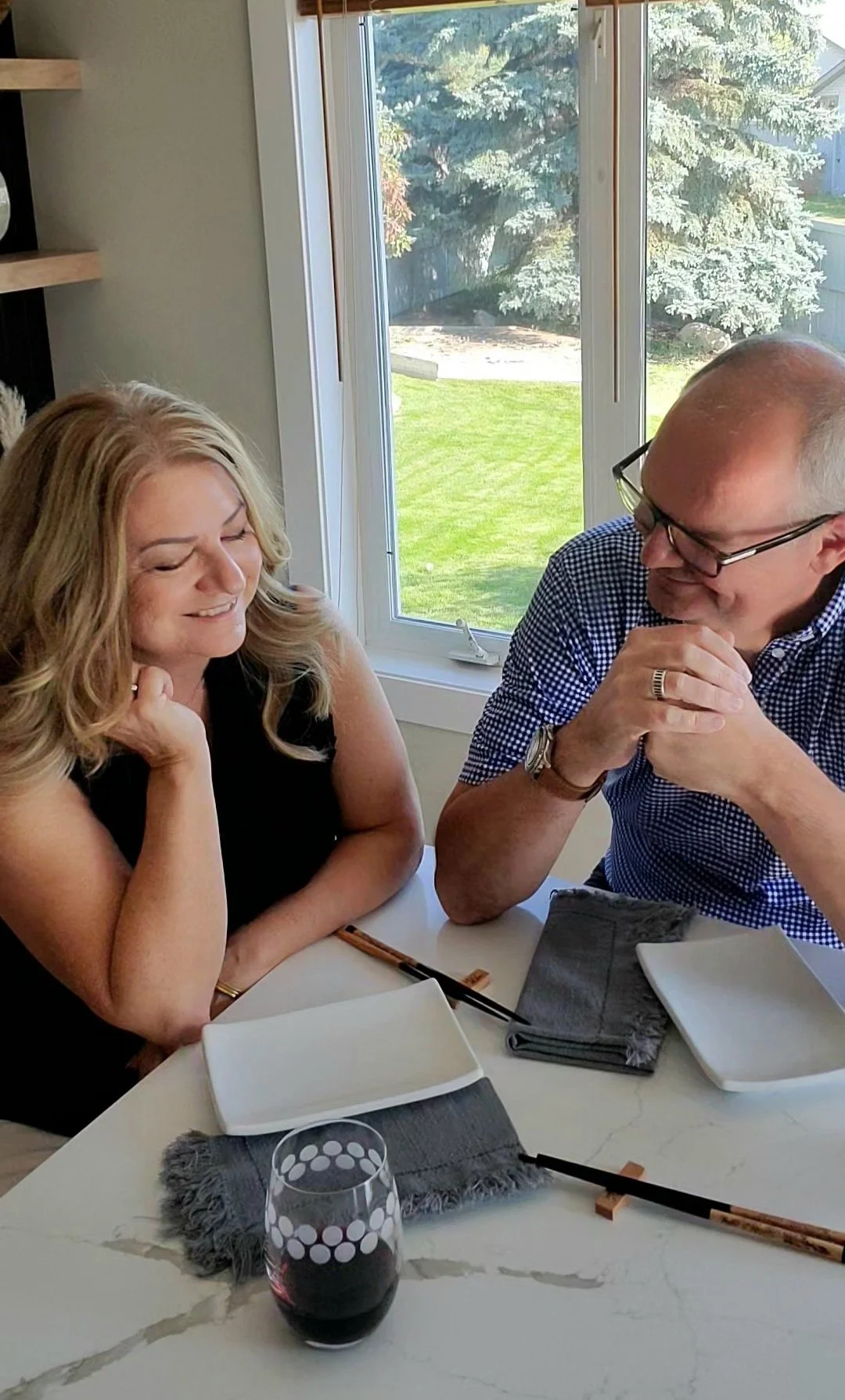 Happy GenX couple laughing together at their kitchen table over dinner with chopsticks and a glass of red wine