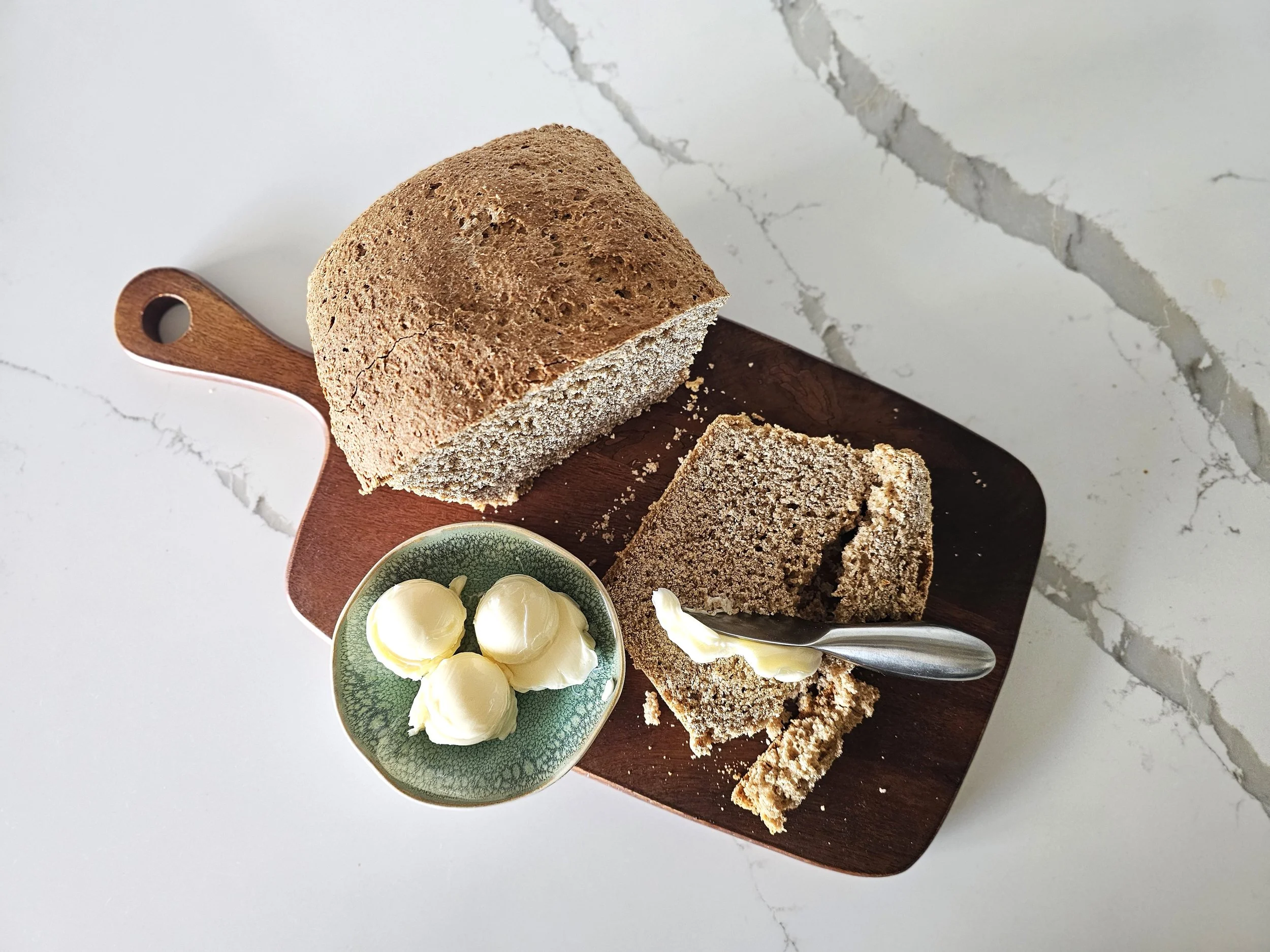 Overhead view of homemade whole wheat bread on a wooden cutting board with butter and a bread knife on a white marble countertop