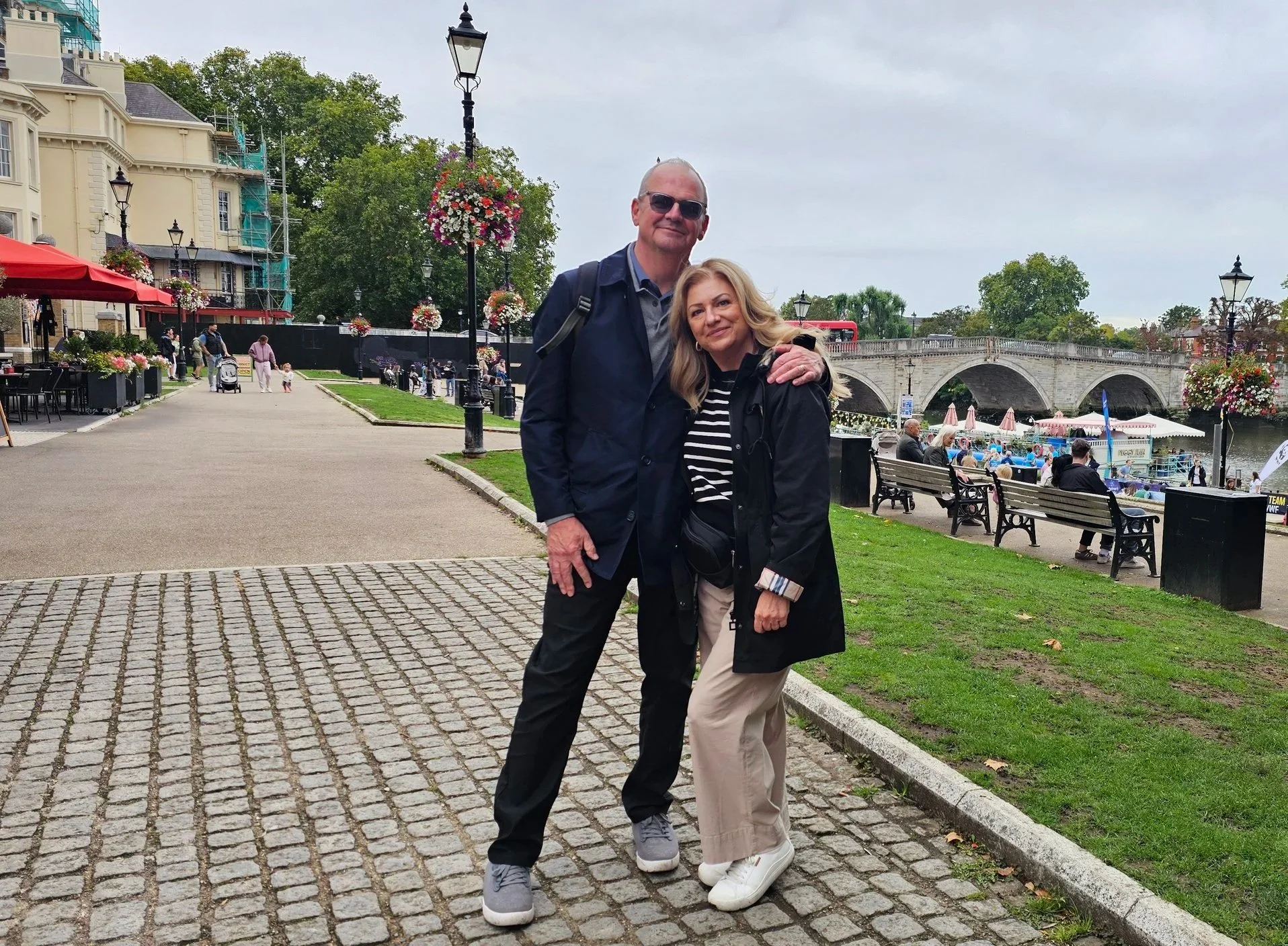 Canadian couple posing together on a cobblestone riverside path in Richmond London with a stone bridge and flower baskets in the background