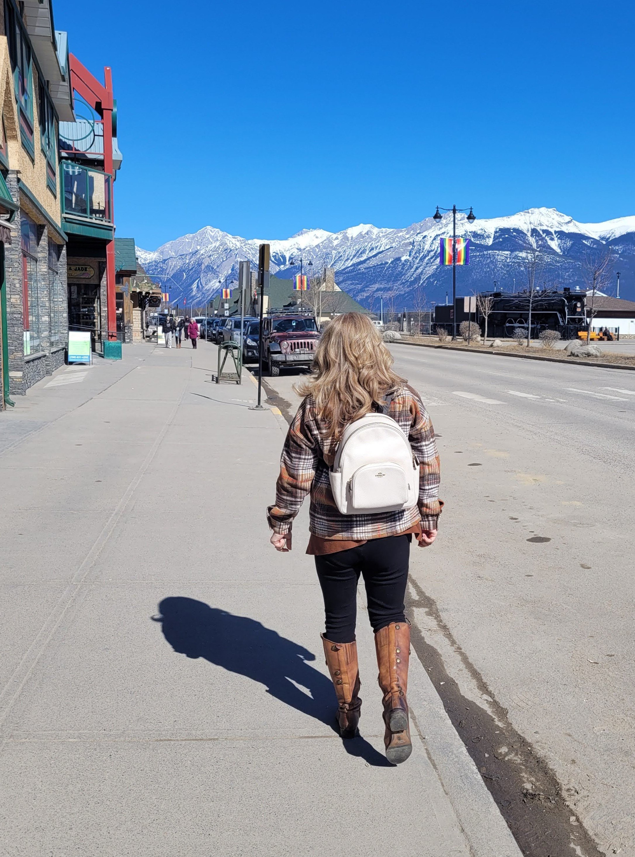 Woman walking along Jasper Alberta main street with white backpack and snow-capped Rocky Mountains under a vivid blue sky