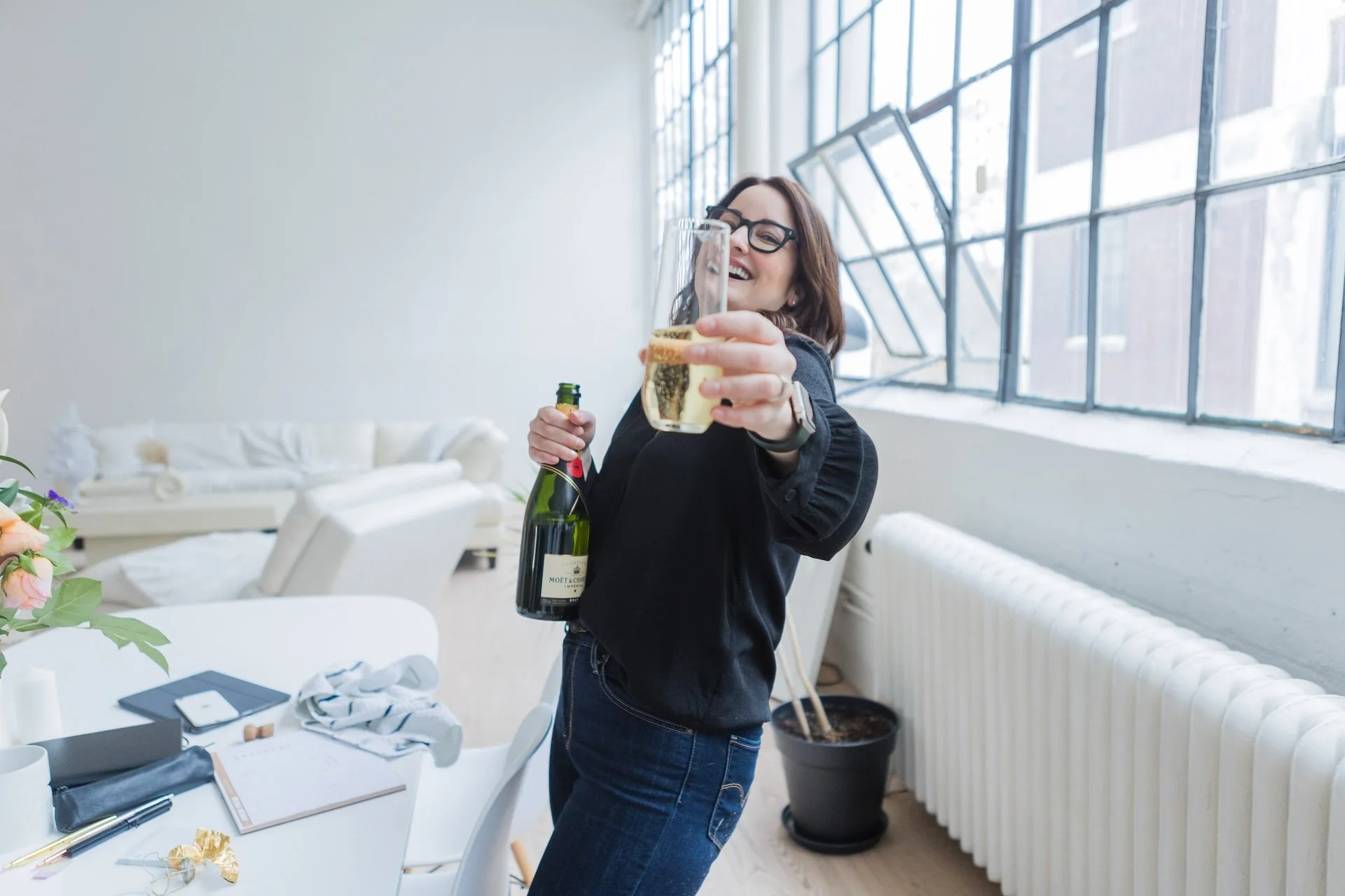 A woman with glasses and brown hair holding a glass of champagne and a bottle, smiling in a bright, modern living room with large windows.