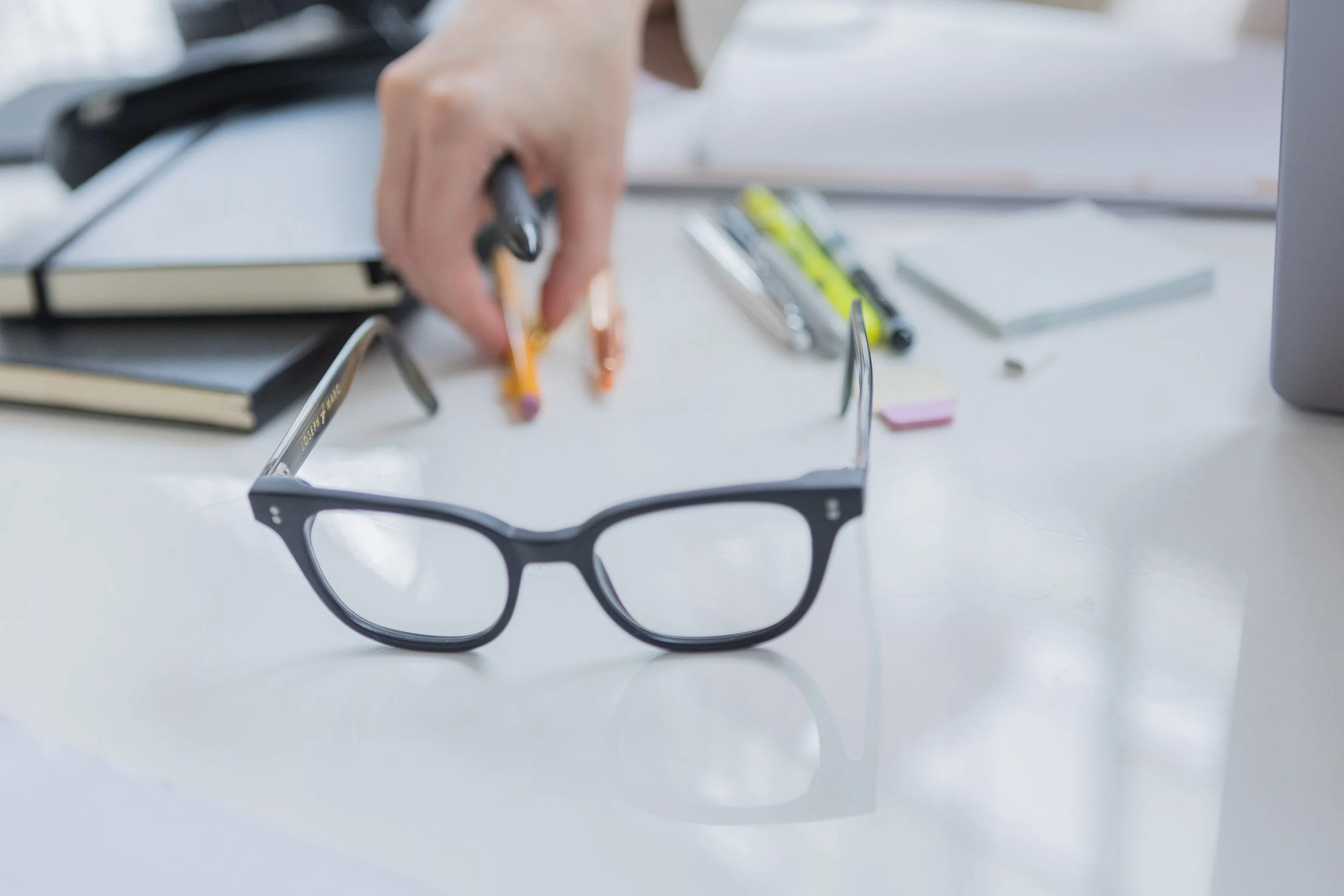 Close-up of black eyeglasses on a white desk with stationery items, hand reaching for pens, and notebooks in the background.