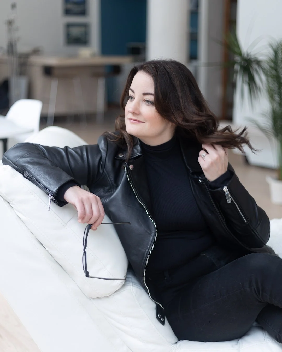 Woman wearing a leather jacket leaning on a chaise playing with her hair