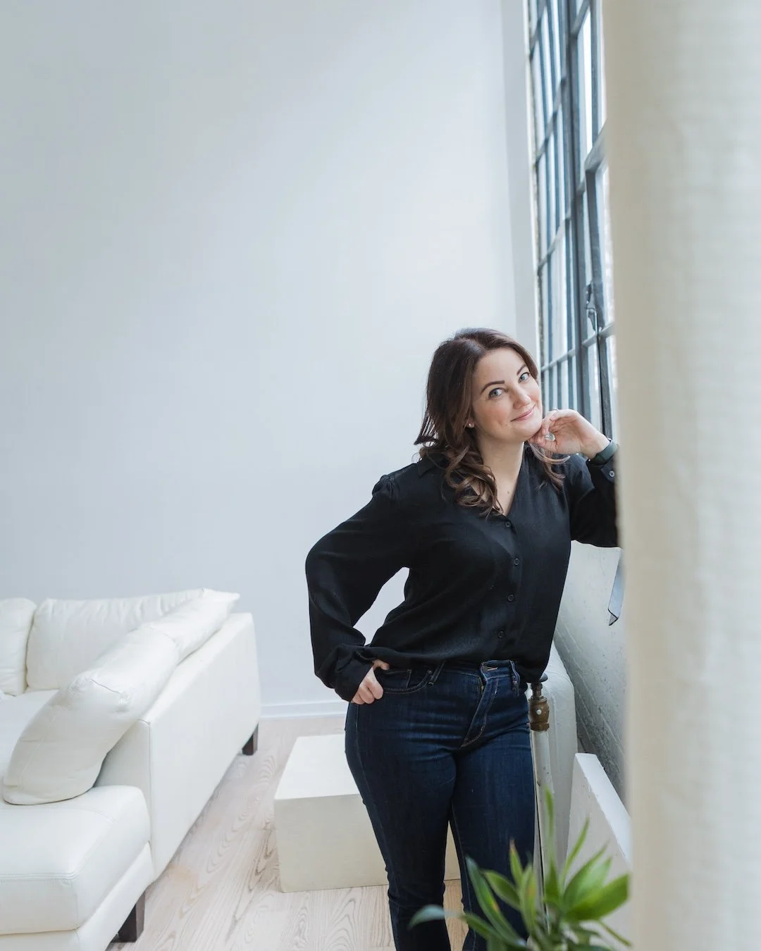 Woman in a black blouse and jeans standing by a large window, white sofa in background.