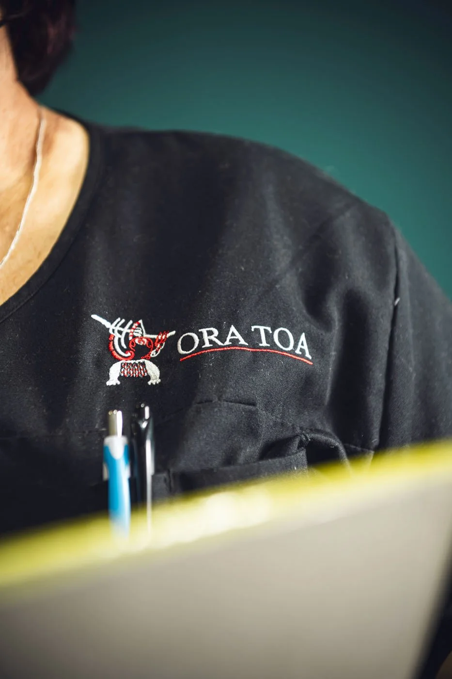 Close-up of a person wearing a black shirt with an embroidered red and white design and the words 'ORA TOA'.