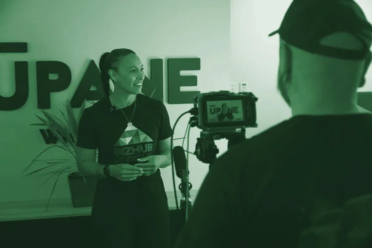 A woman being filmed, standing in front of a yellow wall with large blue letters spelling 'UPHUB'. She is smiling, wearing a black T-shirt with the 'BIZHUB PORI' logo, and has a plant behind her.