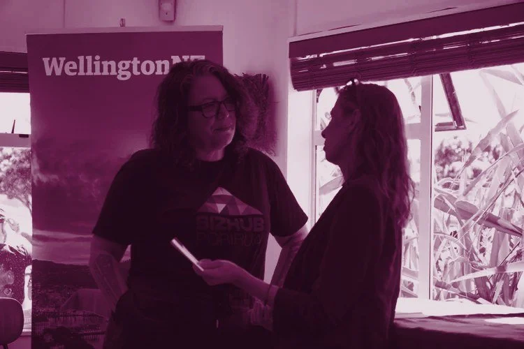 Two women talking indoors near a window with blinds. One woman has curly hair, glasses, tattoos, and is wearing a black T-shirt with a colorful logo and the words 'BIZHUB PORIRUA'. The other woman has long, wavy hair, and is wearing a maroon blazer. Behind them, a Wellington NZ banner is partially visible.