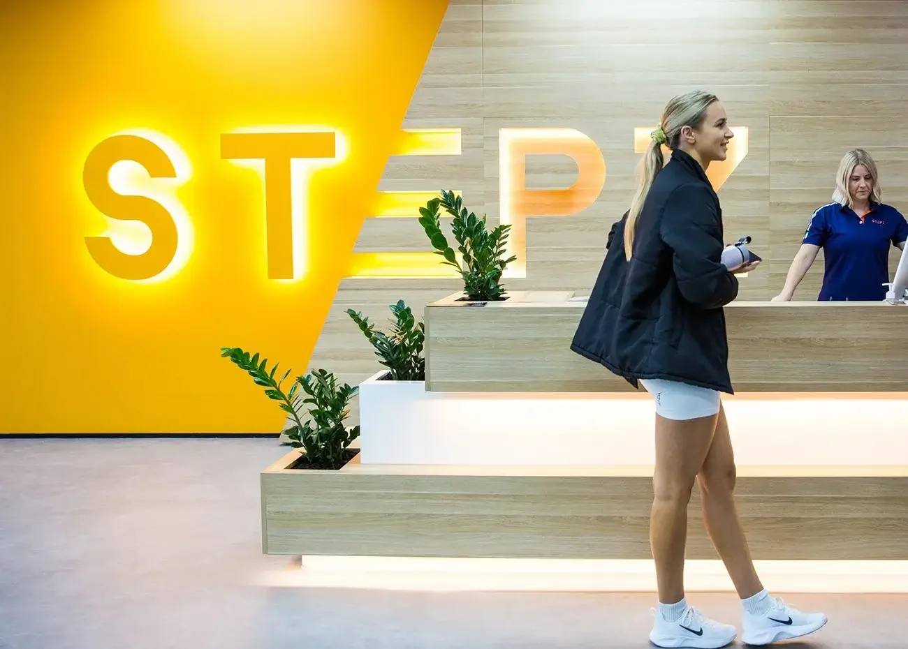 A woman in athletic attire walking past a reception desk in a modern, brightly lit setting with a yellow wall featuring the word 'STEP' in illuminated letters and several potted plants.