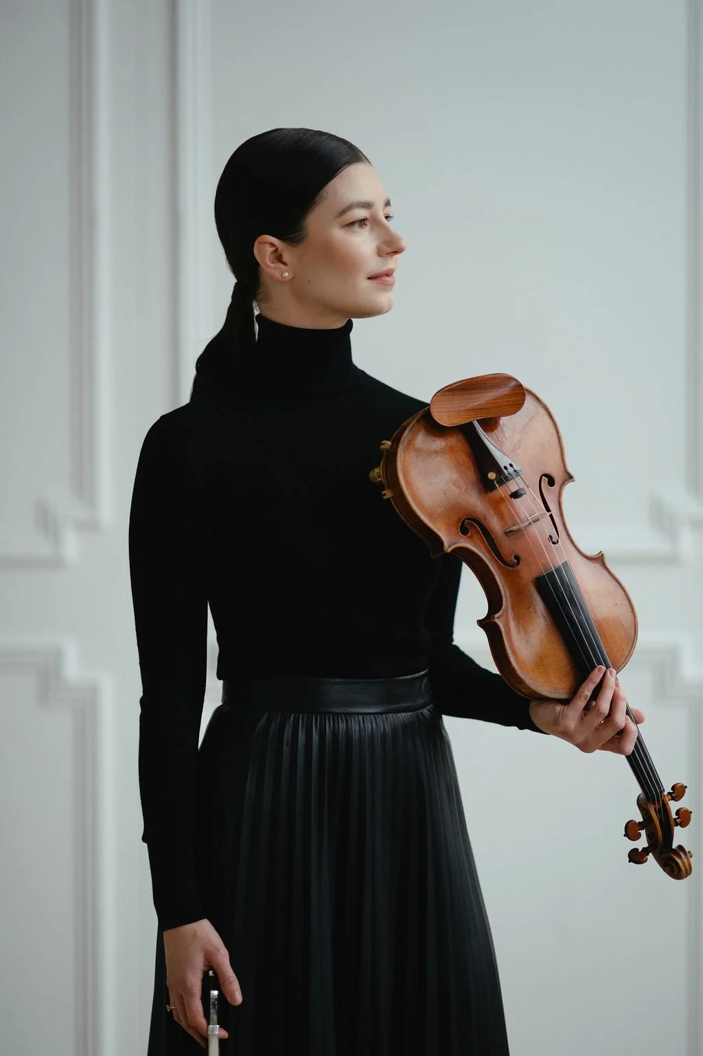 A woman dressed in a black turtleneck and pleated skirt holding a violin in her left hand and a bow in her right hand, standing indoors against a white wall.