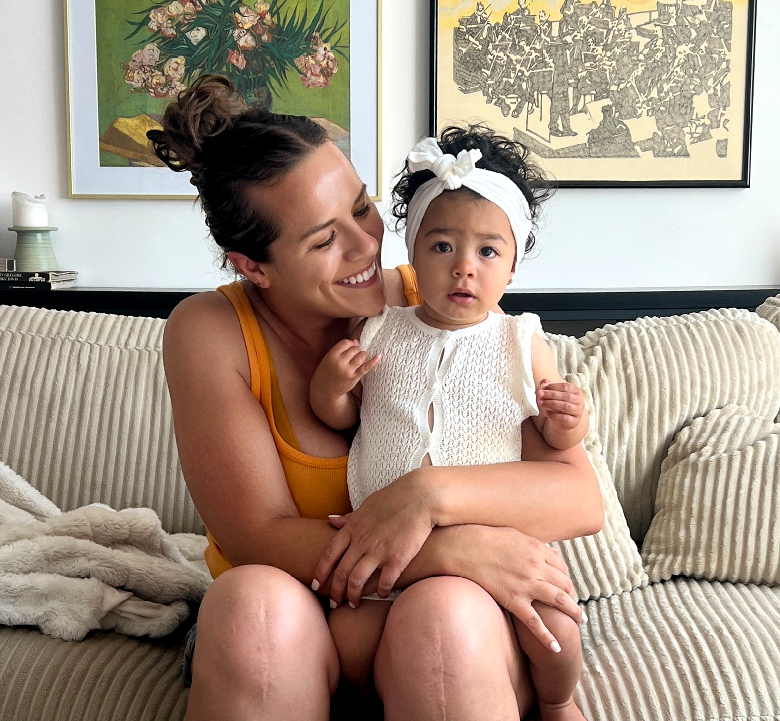 A woman with dark brown hair in a bun holding a young girl with curly hair and a white headband, both sitting on a beige couch in front of a wall with two framed artworks.