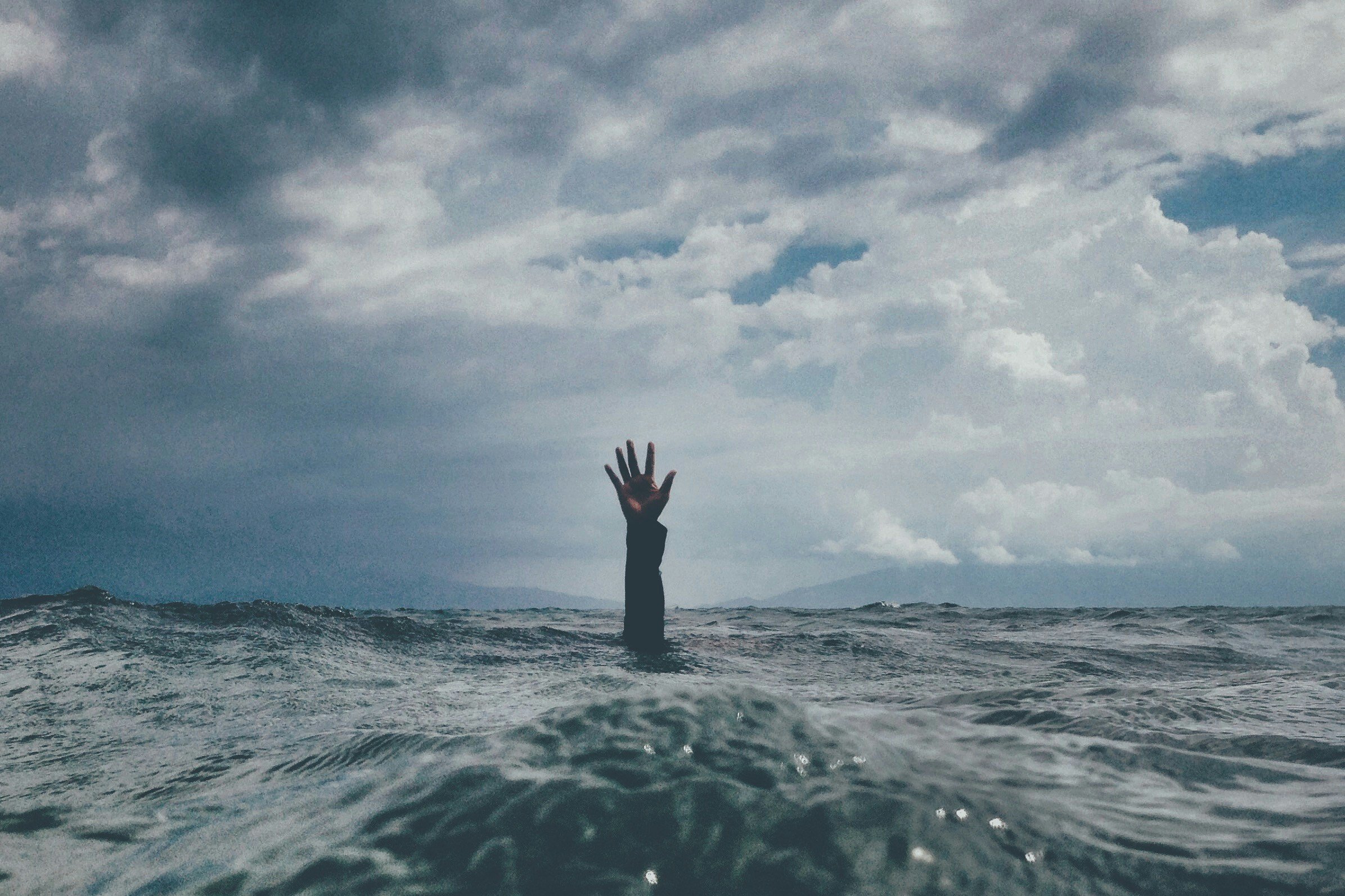 A person’s arm with a black sleeve reaching out of the water in the ocean, with a cloudy sky above.