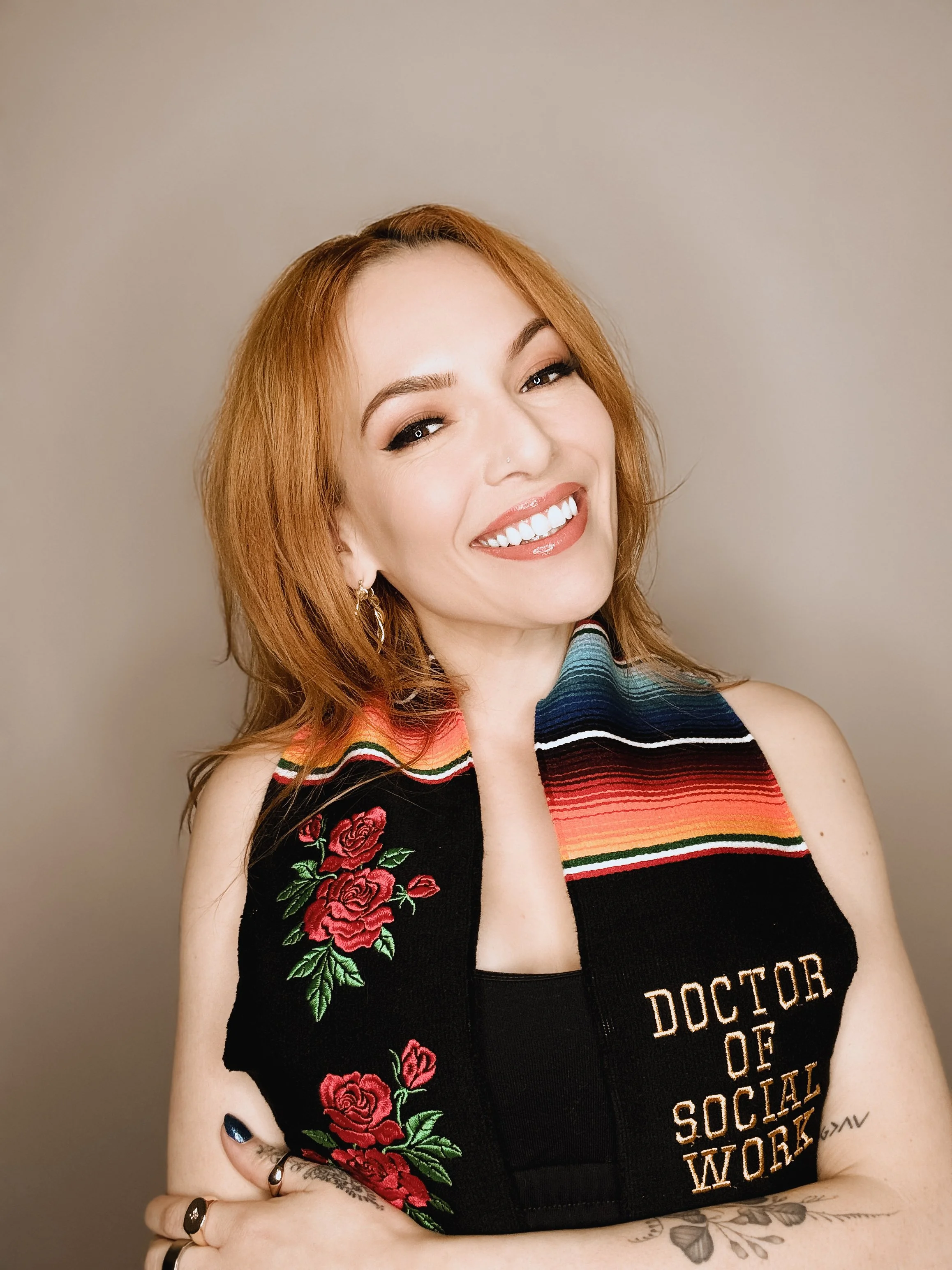 A woman with red hair, wearing a black sleeveless vest with embroidered roses and the words 'Doctor of Social Work,' smiling with crossed arms against a plain background.