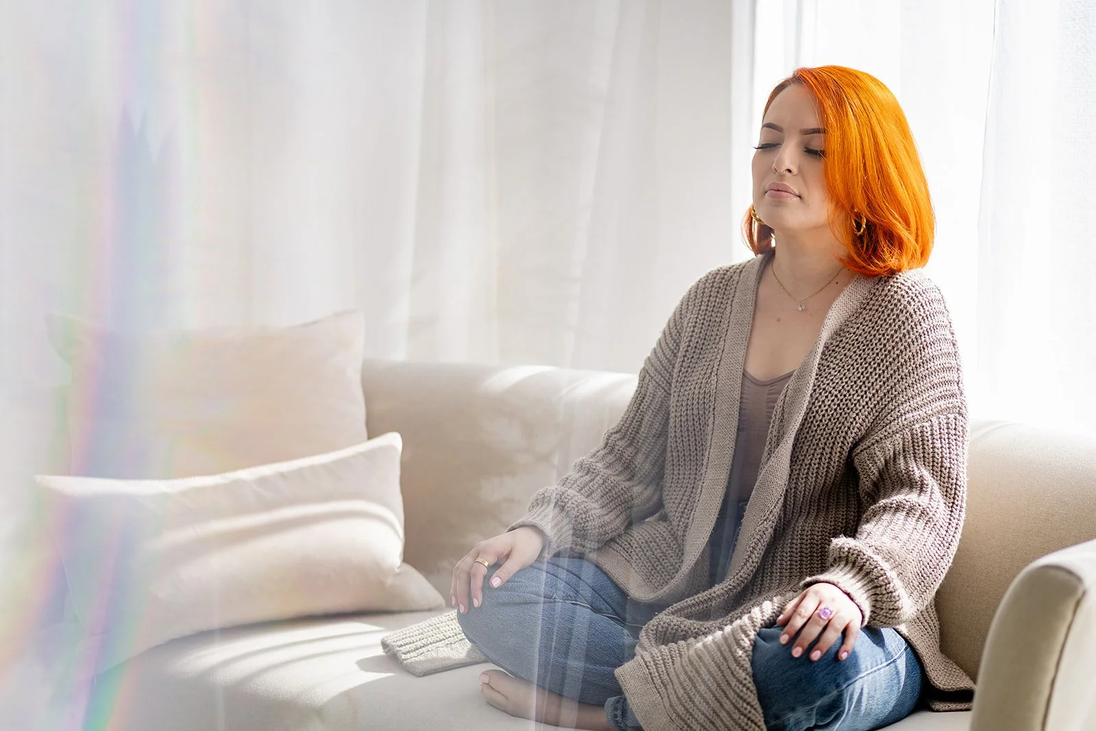 Young woman with vibrant orange hair sitting cross-legged on a beige couch with her eyes closed, meditating in a sunlit living room.