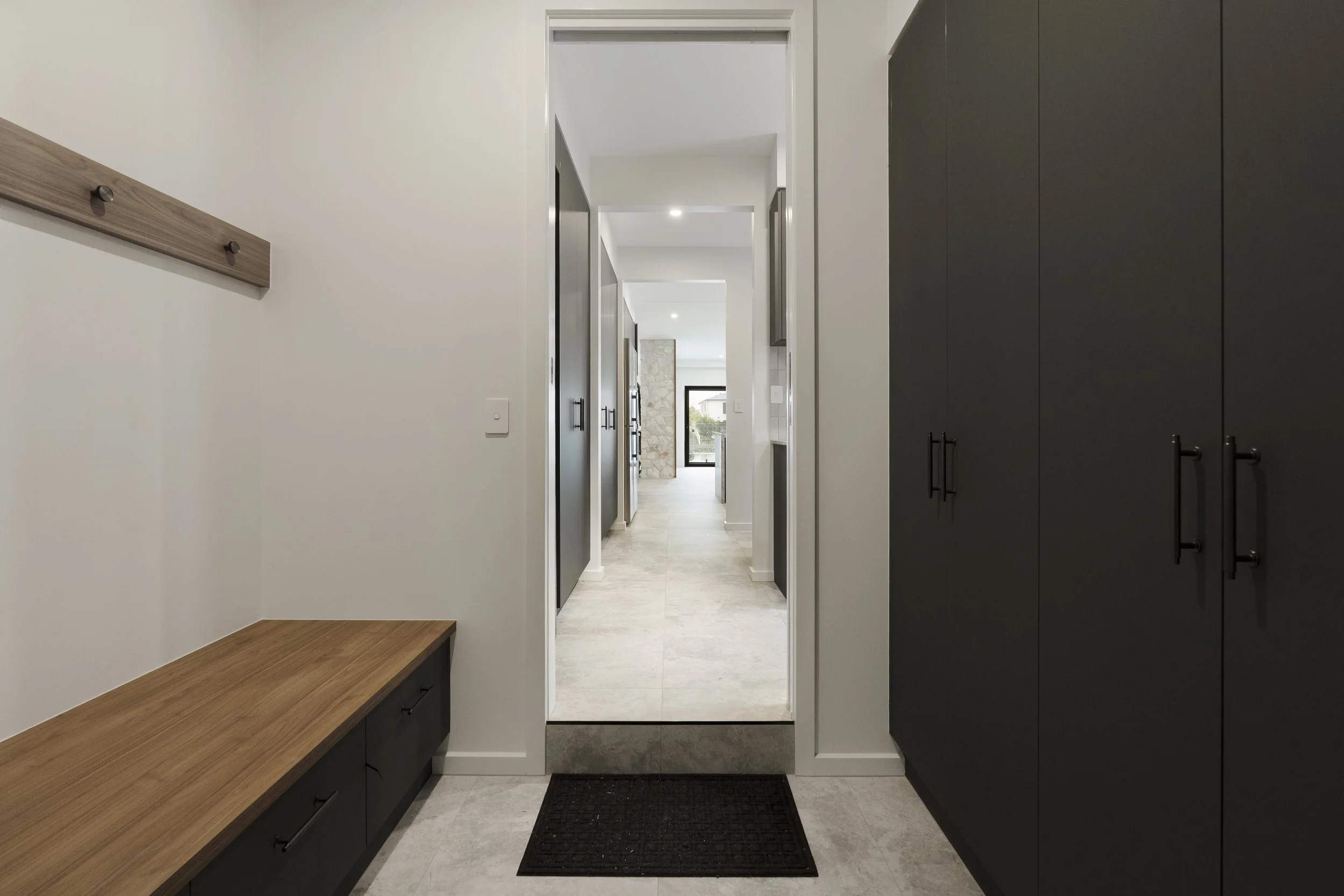 Action Kitchens Interior view of a modern, minimalist hallway with black cabinets on the right, a wooden shelf on the left, and a full-length mirror at the end, leading to a bright living area with large windows.