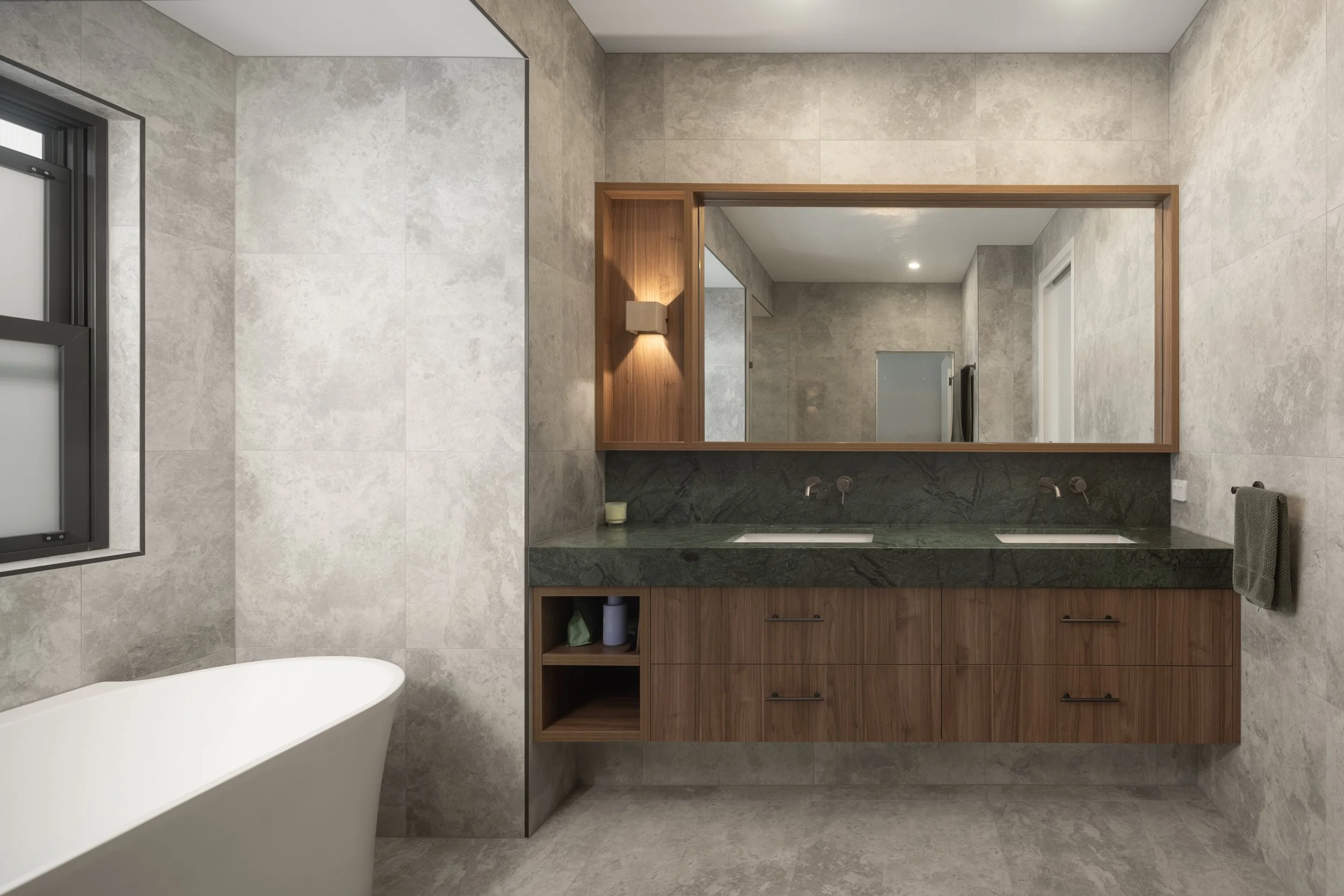 Modern bathroom with a white bathtub, large mirror framed in wood, gray textured tiled walls and floor, and a wooden vanity with two sinks and black drawer handles.  Action Kitchens