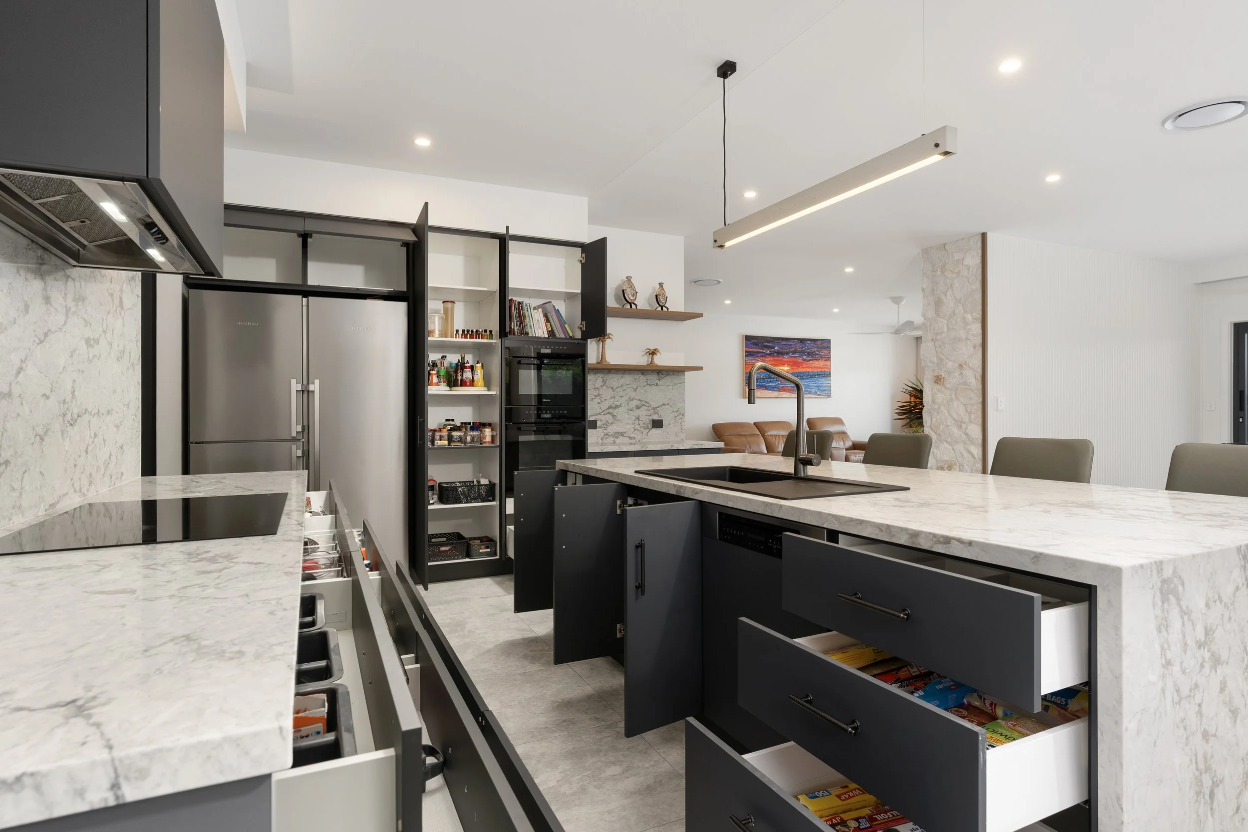 Action Kitchens Modern kitchen with marble countertops, black cabinets, open shelves with spices and books, stainless steel refrigerator, built-in oven, and a view into the living room with a colorful painting, leather seating, and decorative items.