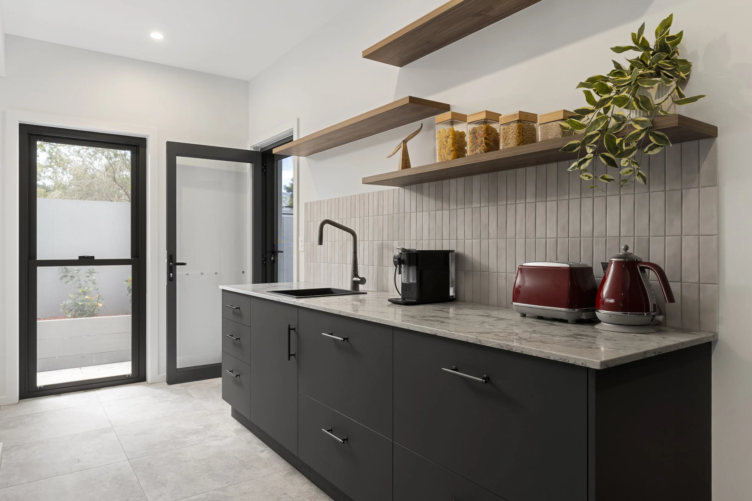 Modern kitchen with gray cabinets, marble countertop, open wooden shelves, and small appliances including a coffee maker and toaster, near a door with a window and outdoor view.