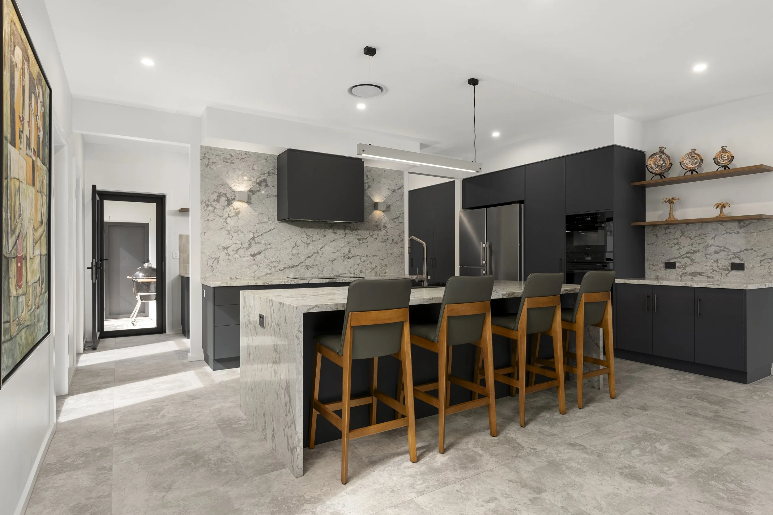Action Kitchens Modern kitchen with white marble island, black cabinets, stainless steel refrigerator, open shelves with decorative vases, and a gray marble accent wall.